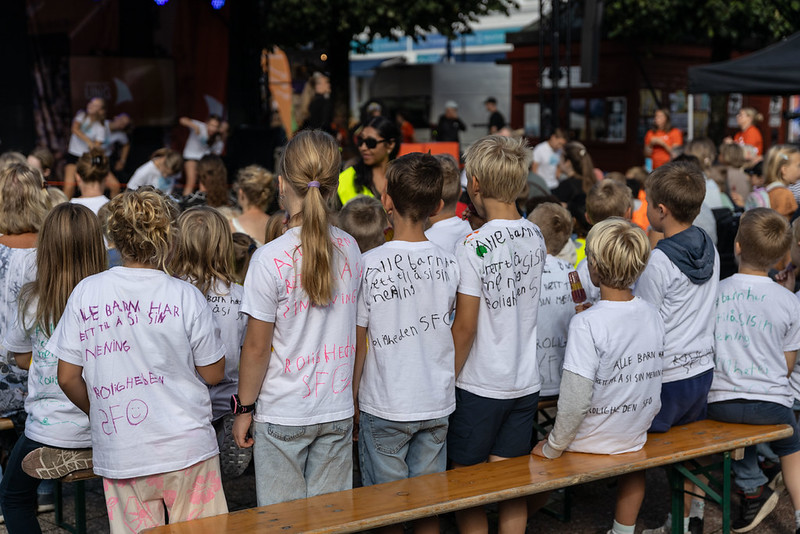 En gruppe barn sitter på en trebenk og ser mot en scene. De har på seg hvite t-skjorter med fargerik skrift og tegninger på ryggen under Arendalsuka Ung.