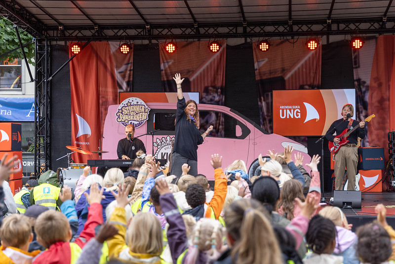 En person står på Torvscenen foran et stort publikum av barn. På scenen står en rosa bil-kulisse, en pianist og en gitarist under Arendalsuka Ung.