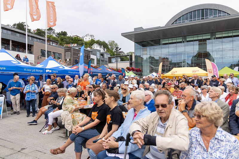 Et stort publikum sitter på benker foran partitelt på et torg under demokratifestivalen Arendalsuka. I bakgrunnen ser man Arendal kultur- og rådhus og flagg som vaier i vinden.