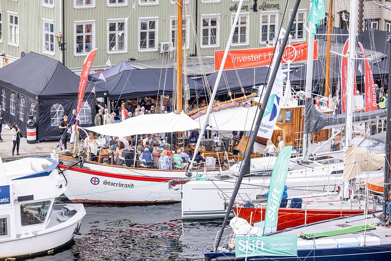 Oversiktsbilde av havnebassenget Pollen fylt med båter. En stor hvit seilskute merket med "Storebrand" har dekket på til gjester under en hvit duk. Rundt ligger flere seilbåter og mindre båter med flagg fra ulike organisasjoner, og i bakgrunnen ser man de karakteristiske trehusene langs brygga.