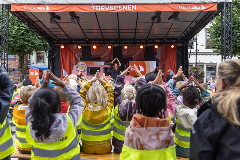 En gruppe barn i gule refleksvester sitter med ryggen til og gjør bevegelser med hendene mot Torvscenen under Arendalsuka ung.