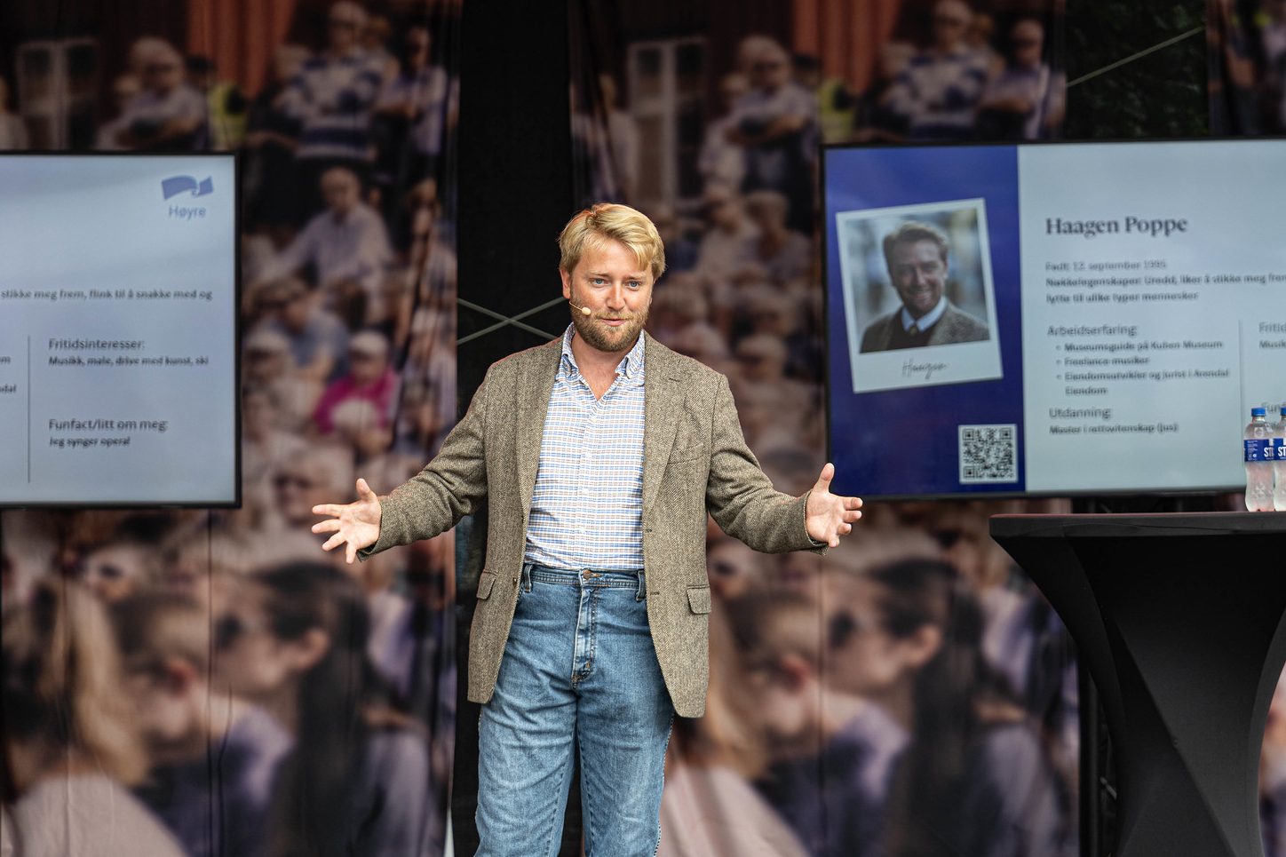 Politiker Haagen Poppe holder et innlegg på en scene under Arendalsuka. Han står mellom to skjermer som viser en presentasjon, kledd i en lys brun blazer og jeans.