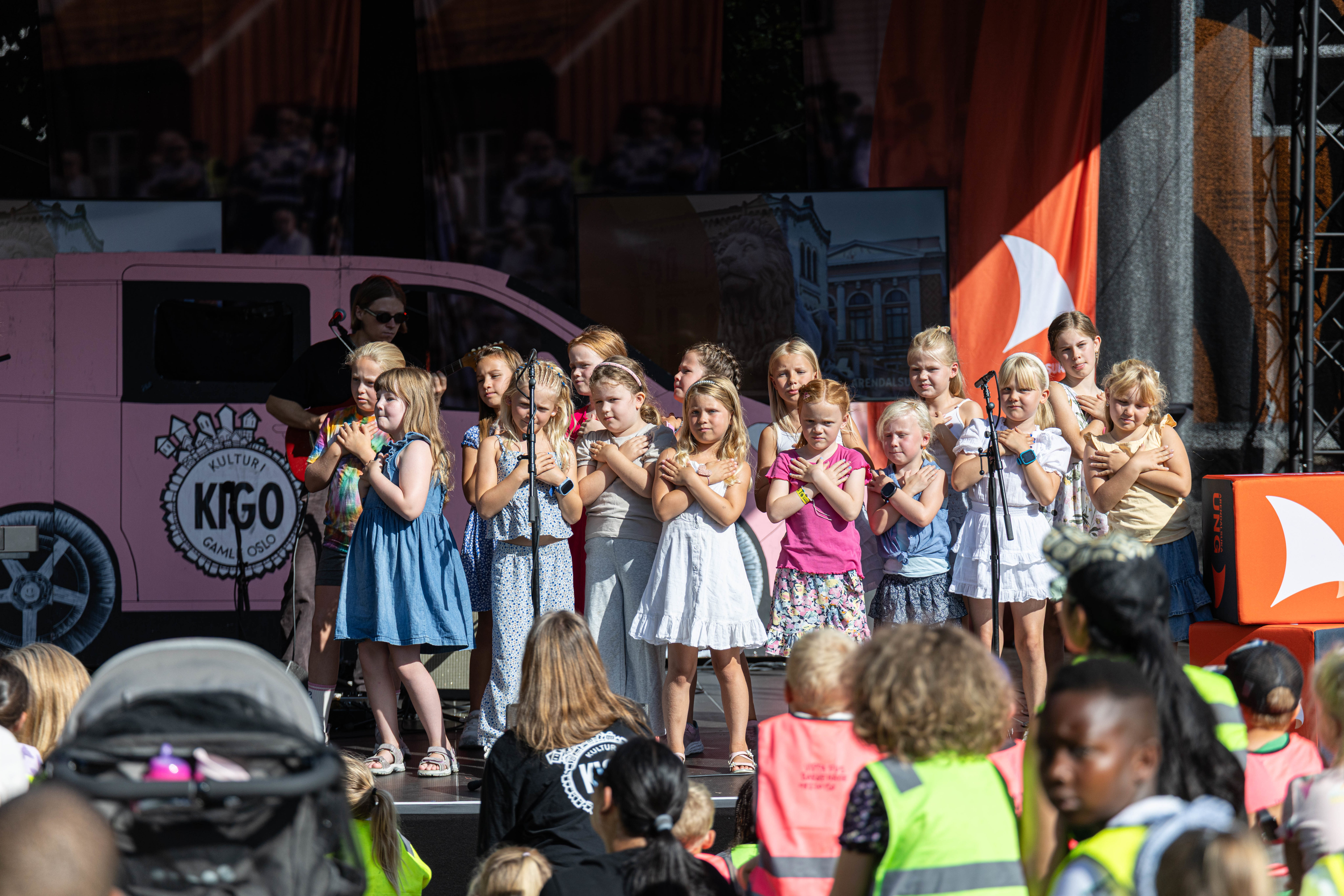 En gruppe på cirka 15 barn står på en utendørsscene og opptrer under Arendalsuka Ung. Barna står i to rekker og holder hendene i kors over brystet mens de synger. Bak dem står en rosa kulisse formet som en bil med logoen "KIGO – Kultur i Gamle Oslo". I forgrunnen ser man bakhodet til et publikum, inkludert flere barn i fargerike refleksvester som følger med på forestillingen i solskinnet.