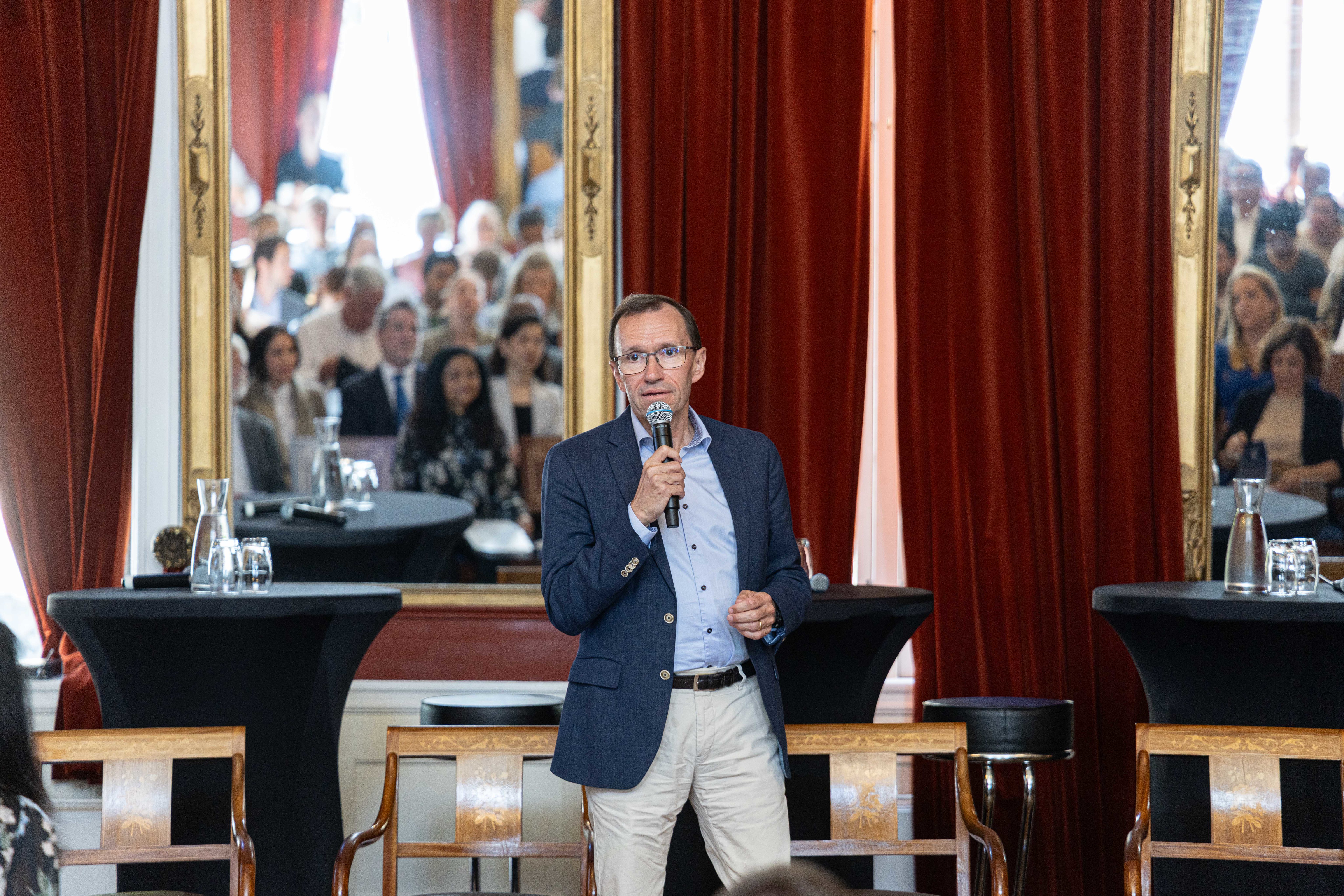 En mann med briller, iført blå dressjakke og lyse bukser, taler i en mikrofon i et elegant rom med historisk preg. Han står foran to store speil med gullrammer som reflekterer et lyttende publikum. Rommet er dekorert med tunge, rødbrune gardiner og klassiske trestoler med mønstret trekk. Foran ham står svarte kafébord med vannkarafler og glass, noe som skaper en formell, men intim ramme for foredraget.