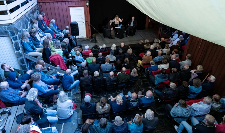 Et tettpakket publikum sitter i en intim bakgård med vegger i tre og stein. På en liten scene sitter tre personer i en samtale under en lys duk som skjermer for solen.