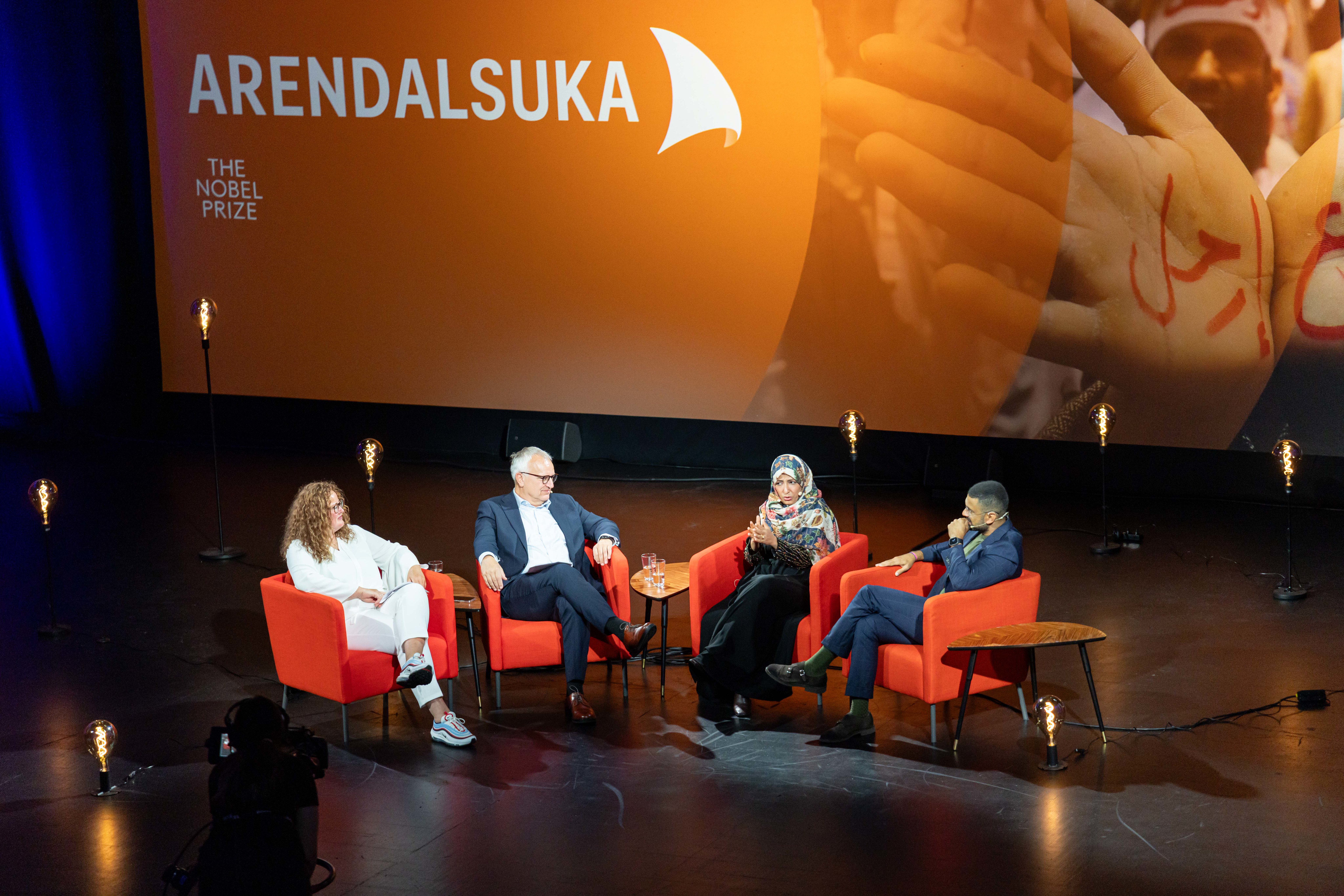 Fire personer sitter i røde lenestoler på en mørk scene under et arrangement for Arendalsuka. Bak dem er en stor oransje skjerm med logoene til "Arendalsuka" og "The Nobel Prize". En kvinne i midten snakker engasjert med hendene, mens de tre andre lytter. Scenen er dekorert med flere glødepærer på lange stenger som står spredt rundt stolene, og et stort bilde av hender projiseres på den høyre delen av bakskjermen.
