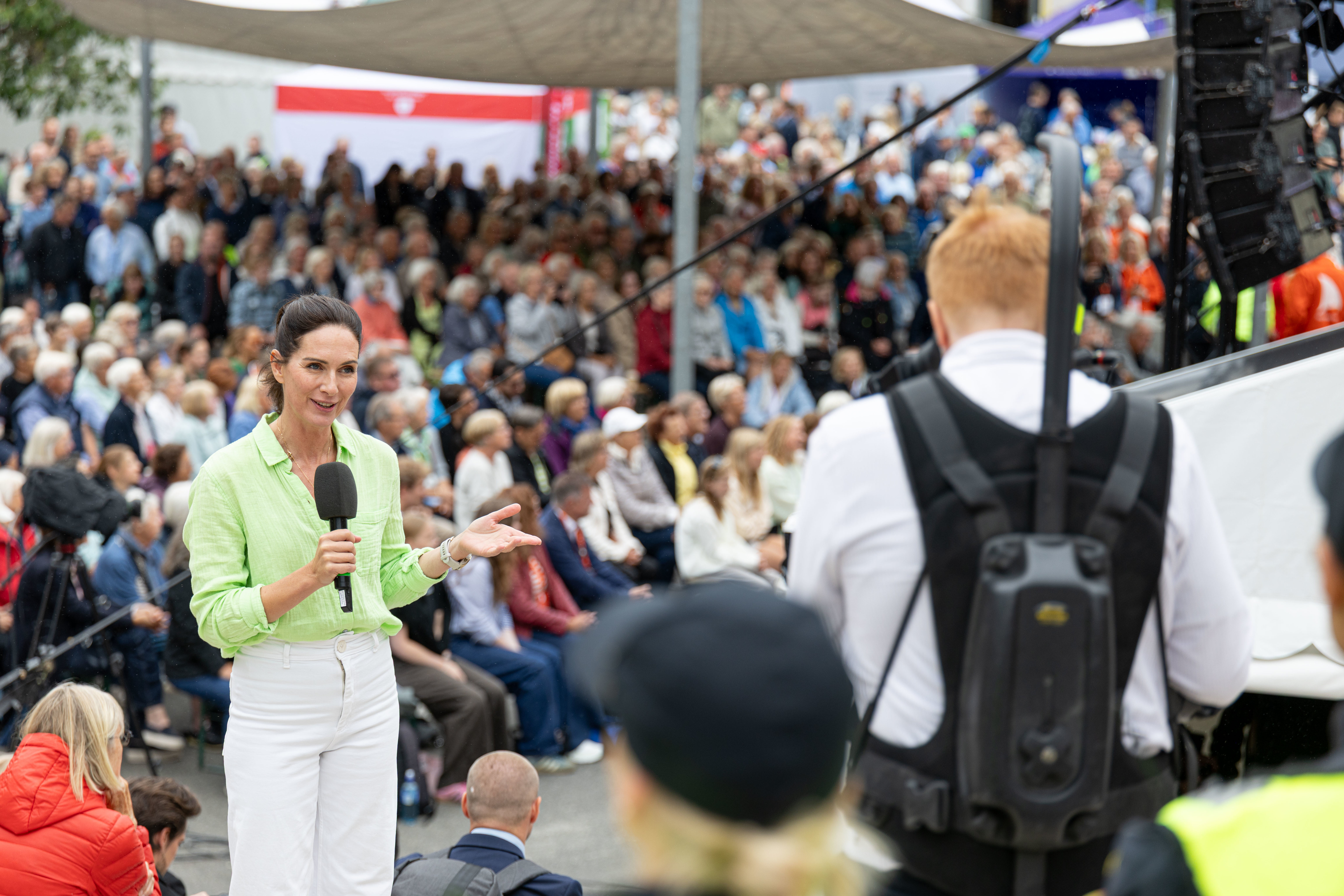 En kvinne i lysegrønn skjorte og hvit bukse snakker inn i en mikrofon foran en stor folkemengde utendørs under Arendalsuka. Hun gestikulerer med den ene hånden mens hun holder mikrofonen i den andre. I forgrunnen ser vi ryggen til en fotograf med et avansert kamerasystem festet til en bæresele. Bakgrunnen er fylt med et sittende publikum under en stor, lys seilduk som skjermer for solen, og fargerike telt og flagg kan skimtes i det fjerne.
