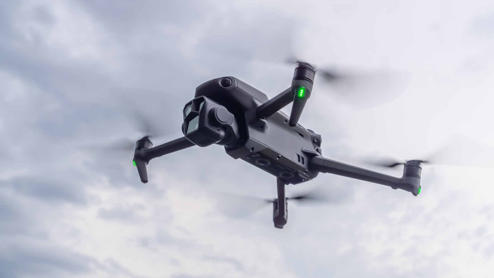 En mørkegrå drone flyr i luften mot en overskyet, lys grå himmel. Dronens fire propeller er i rask bevegelse og fremstår som uskarpe sirkler. Små, grønne lys lyser på undersiden av hver motorarm, og det avanserte kameraet foran på dronen er rettet skrått nedover. Bildet er tatt nedenfra, noe som gir et tydelig perspektiv av dronens underside og teknologi.