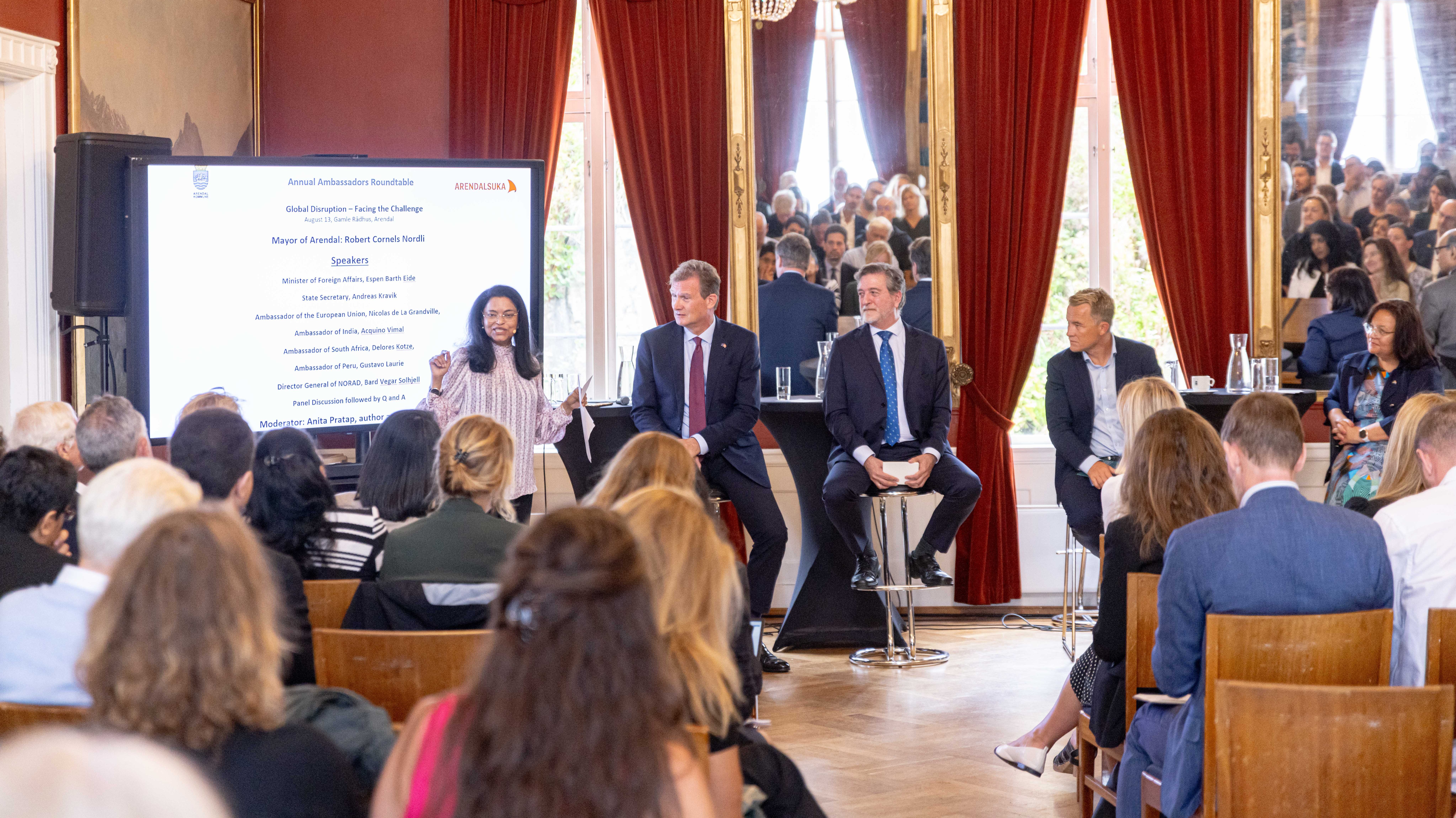 En kvinne står og taler ved siden av en stor skjerm under et internasjonalt arrangement i et staselig rom med røde vegger og gullforgylling. Tre menn i mørke dresser sitter på barkrakker i panelet ved siden av henne, og i bakgrunnen ser man et lyttende publikum. Skjermen viser programmet for «Annual Ambassadors Roundtable» med temaet «Global Disruption – Facing the Challenge». Rommet er preget av klassisk arkitektur med store speil og tunge, røde gardiner som rammer inn vinduene.