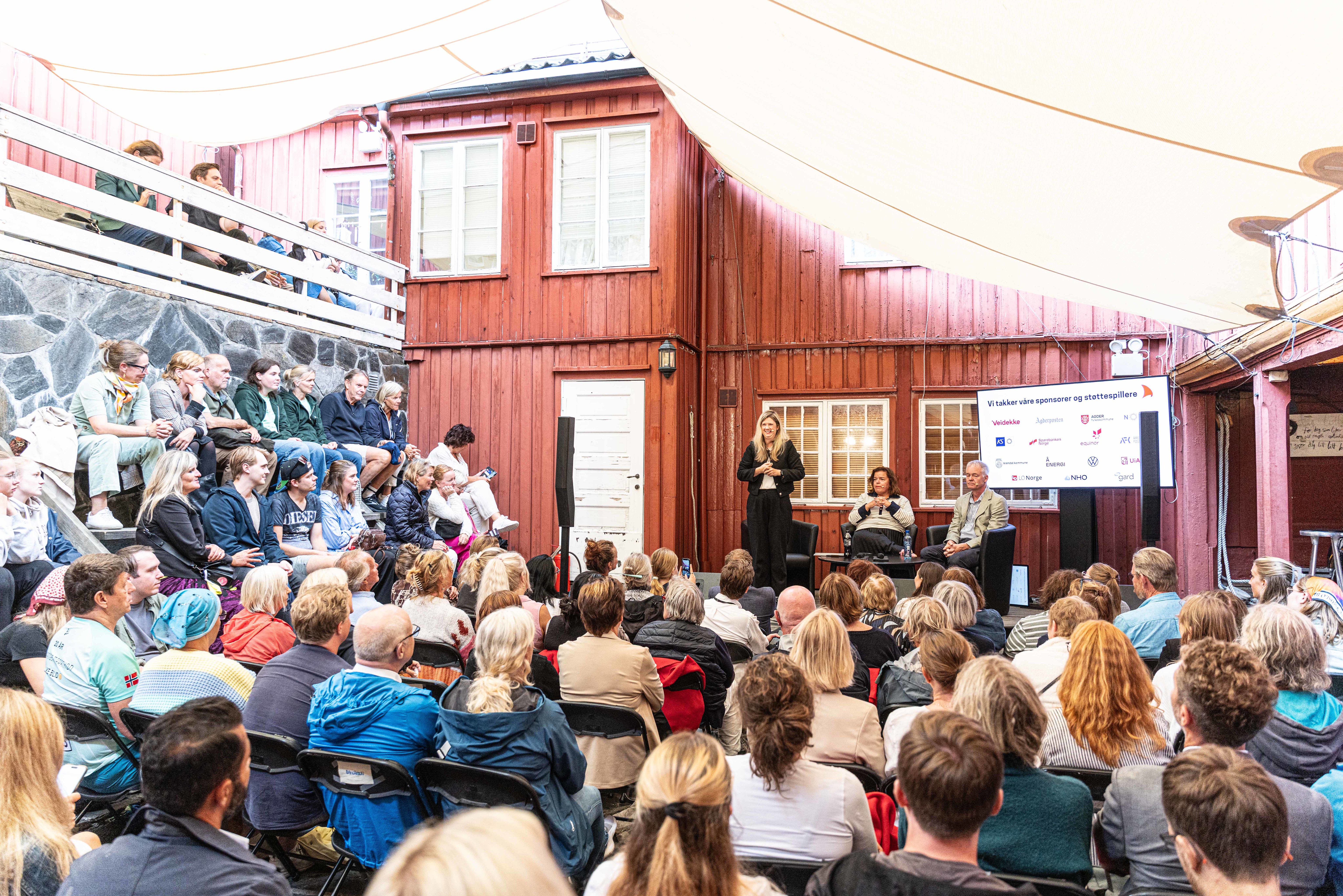 En fullsatt bakgård under et arrangement i Arendalsuka. Et stort publikum sitter på stoler og i en steintrapp og ser mot en liten scene foran en rød trebygning. På scenen står en kvinne og snakker ved siden av to andre personer som sitter i stoler. Ved siden av dem står en skjerm med teksten "Vi takker våre sponsorer og støttespillere" og ulike logoer. En lys solskjerming er spent ut over hele bakgården og skaper et lunt uterom.