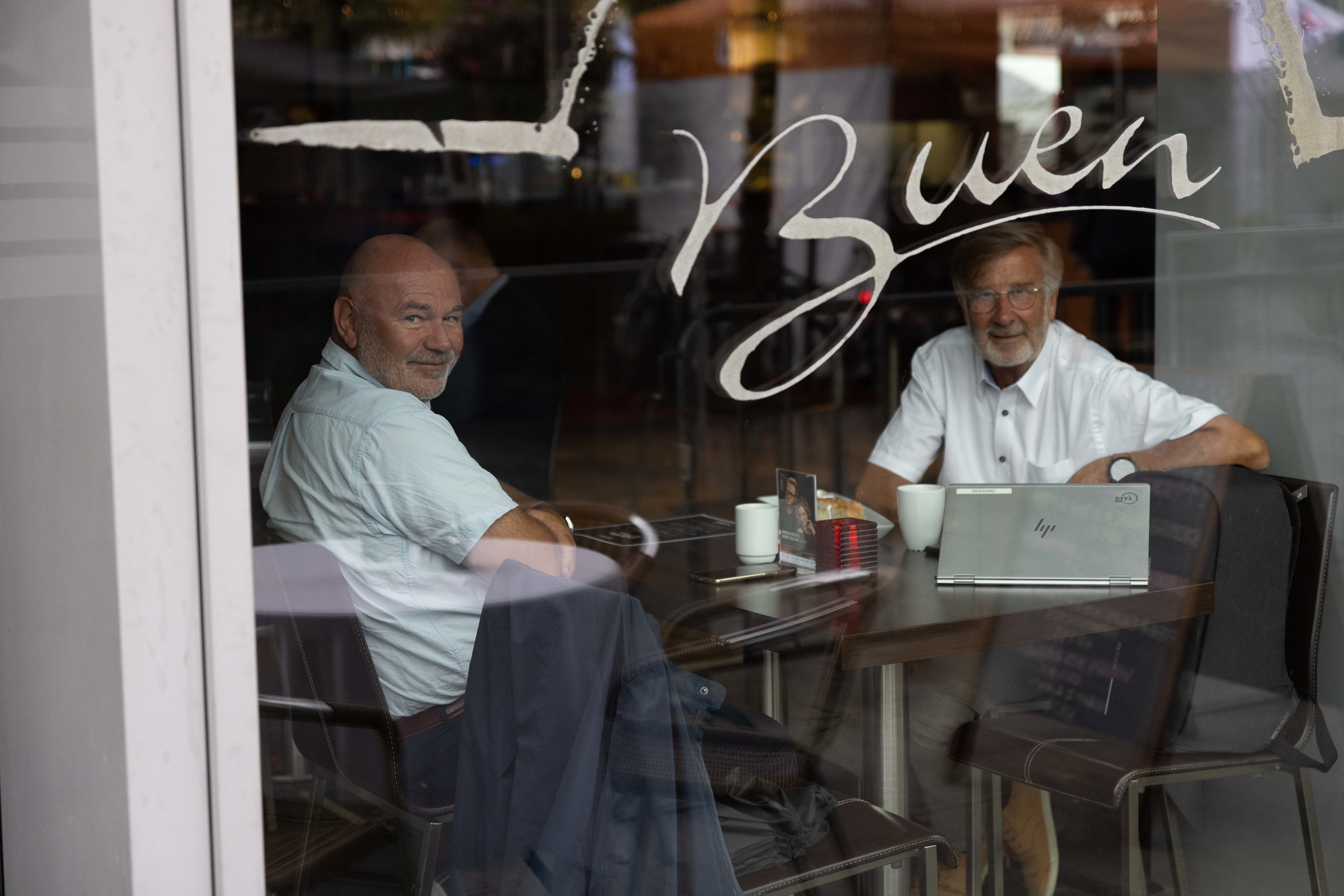 To menn sitter ved et bord inne i et lokale, sett gjennom en glassrute med påskriften "Buen". De sitter overfor hverandre med en bærbar PC, mobiltelefoner og kaffekopper på bordet. Begge ser smilende mot kamera. Refleksjoner i glasset viser et utendørsområde, noe som skaper en dybdeeffekt mellom personene inne og omgivelsene utenfor.