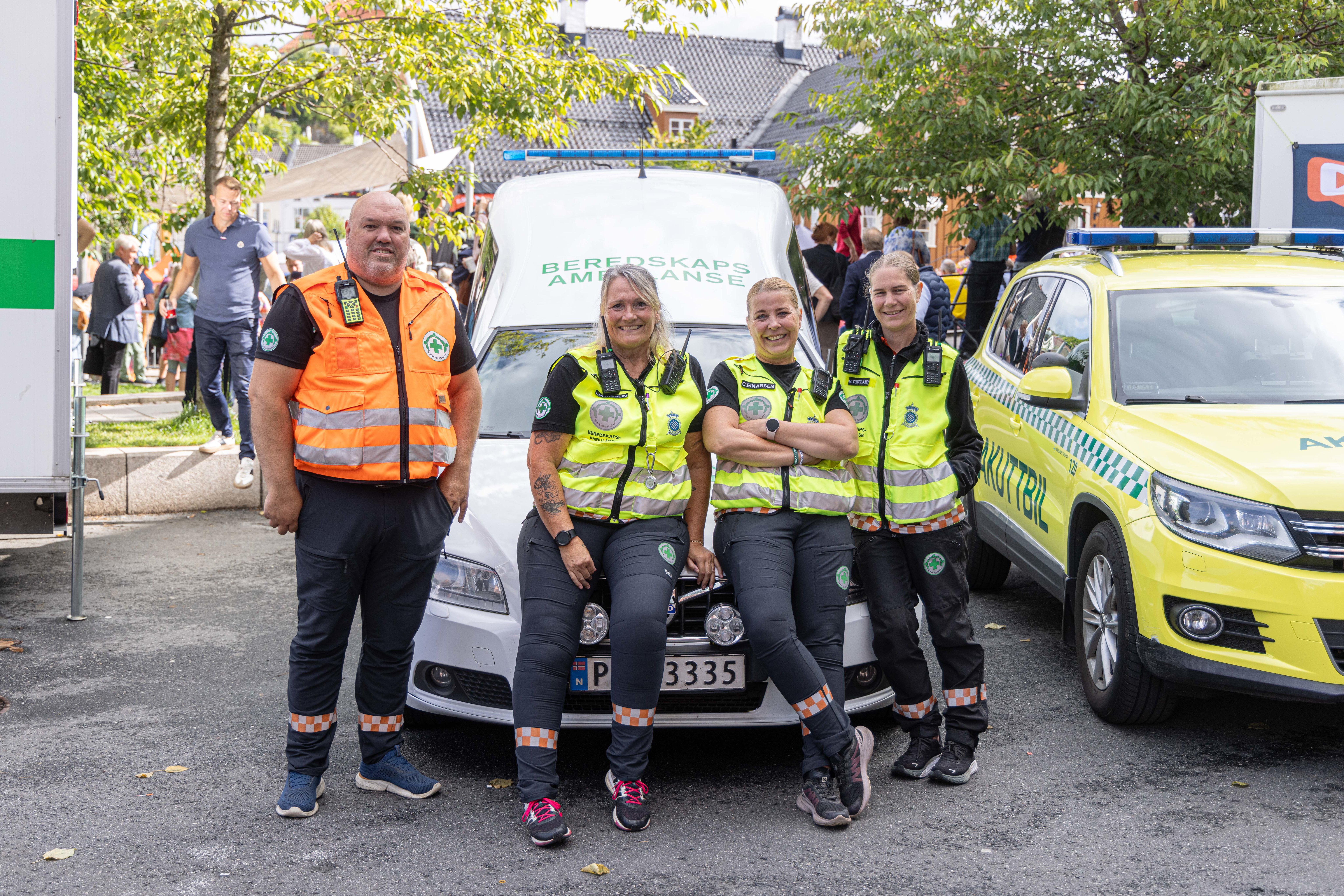 Fire smilende personer fra beredskapstjenesten poserer foran en hvit ambulanse merket "Beredskapsambulanse" og en gul akuttbil. Til venstre står en mann i oransje refleksvest, mens tre kvinner i gule reflekssvester sitter på panseret og står ved siden av ambulansen. Alle bærer profesjonelt radioutstyr og uniformer med organisasjonens logo. Bak dem er det grønne trær og folk som beveger seg i et solfylt uteområde under Arendalsuka.