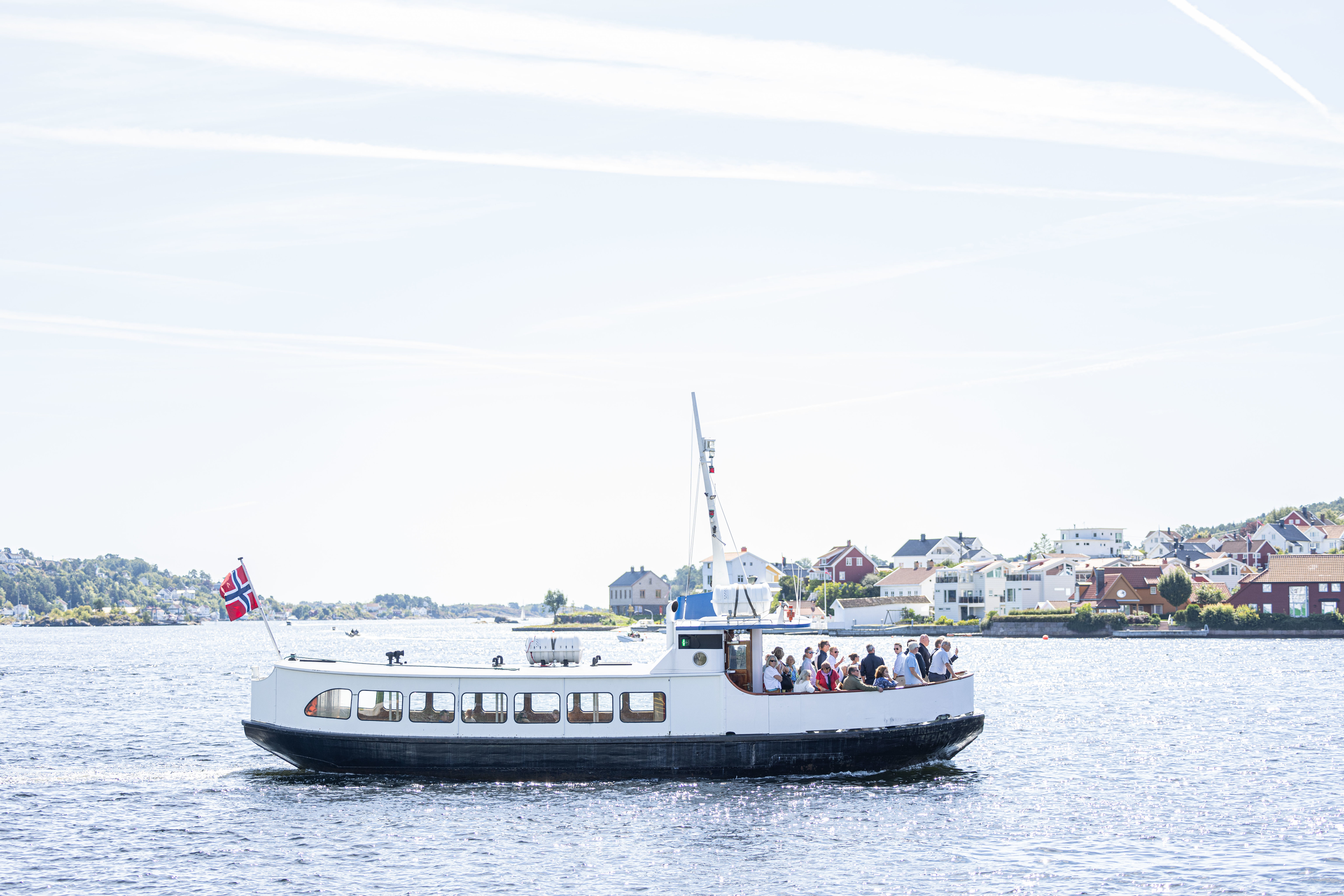 En hvit og svart passasjerferge, "Kolbjørn", kjører over et glitrende blått vann i strålende solskinn. Fergen har et stort norsk flagg akterut, og dekket er fylt med passasjerer som sitter utendørs og nyter turen. I bakgrunnen ser man den idylliske kystlinjen med hvite og røde trehus som ligger tett i tett langs vannkanten under en lys himmel med tynne skystriper.