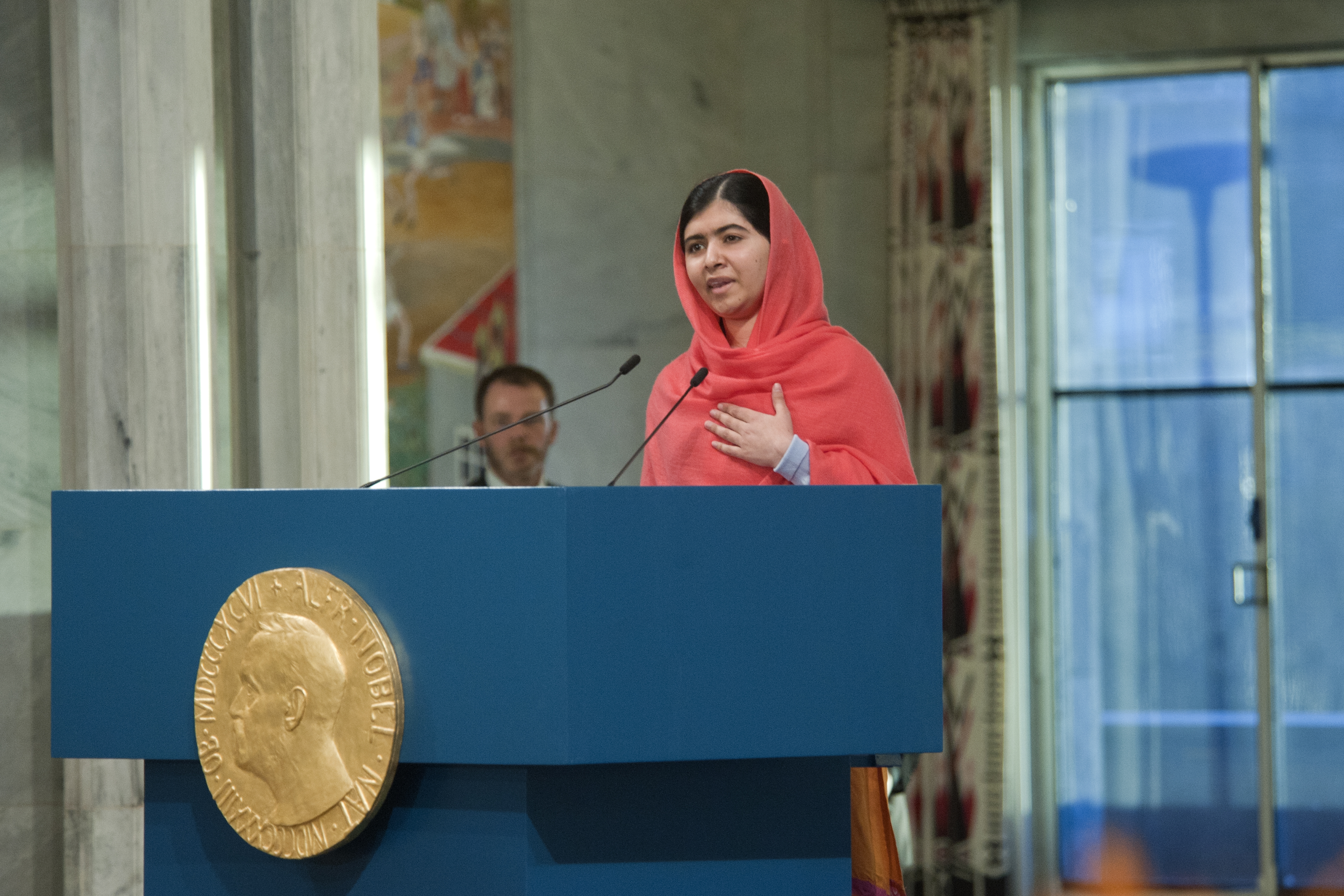 Malala Yousafzai holder tale ved en blå talerstol dekorert med Nobels fredspris-medaljen.