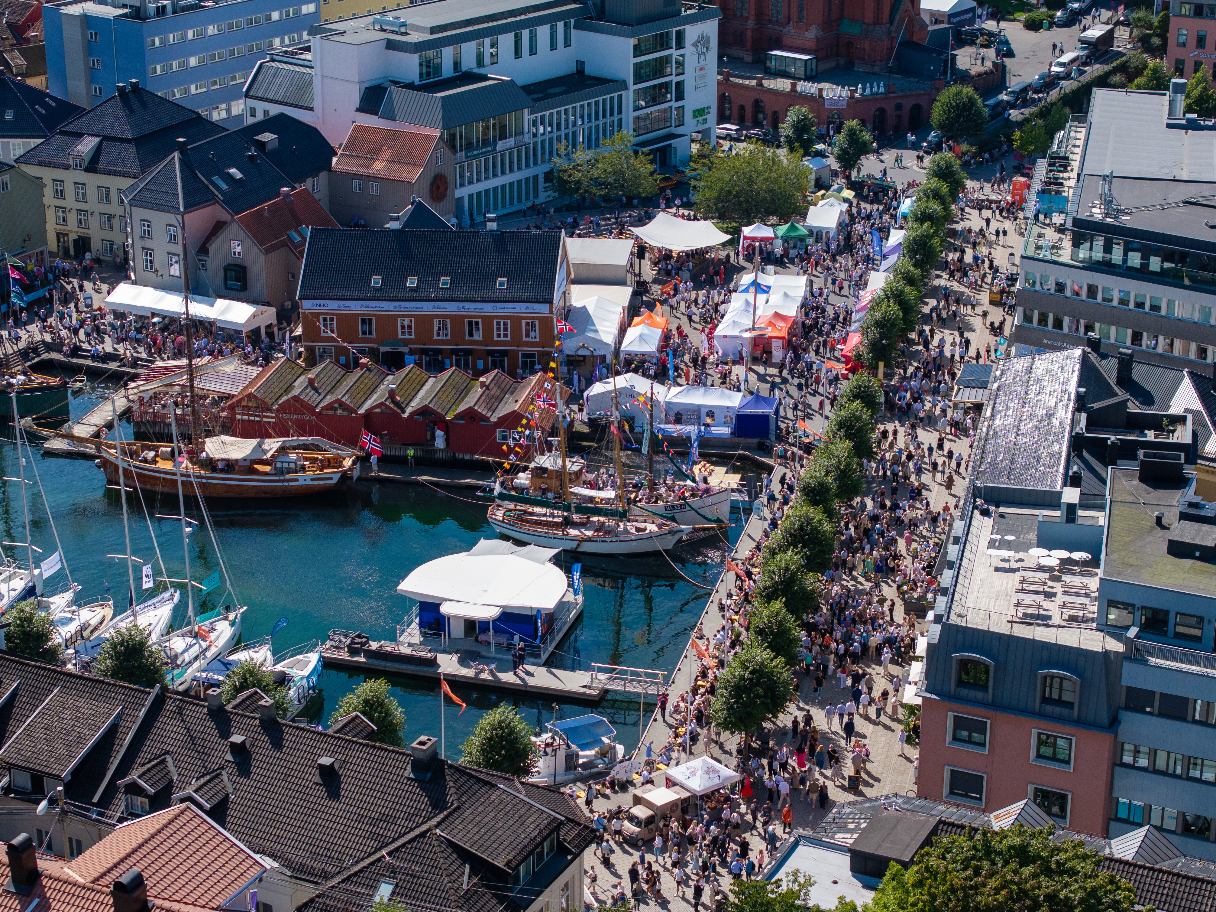 Et oversiktsbilde tatt fra høyden over Pollen i Arendal under Arendalsuka. Havnebassenget er fylt med båter, inkludert store tradisjonelle seilskuter og moderne seilbåter. Langs kaikanten yrer det av en tettpakket folkemengde som beveger seg mellom en mengde hvite og fargerike telt, boder og stands. Arkitekturen i bakgrunnen er en blanding av historiske trehus i gult, oransje og rødt, og moderne kontorbygg i glass og betong. Det er en solfylt dag med klart blått vann og god stemning.