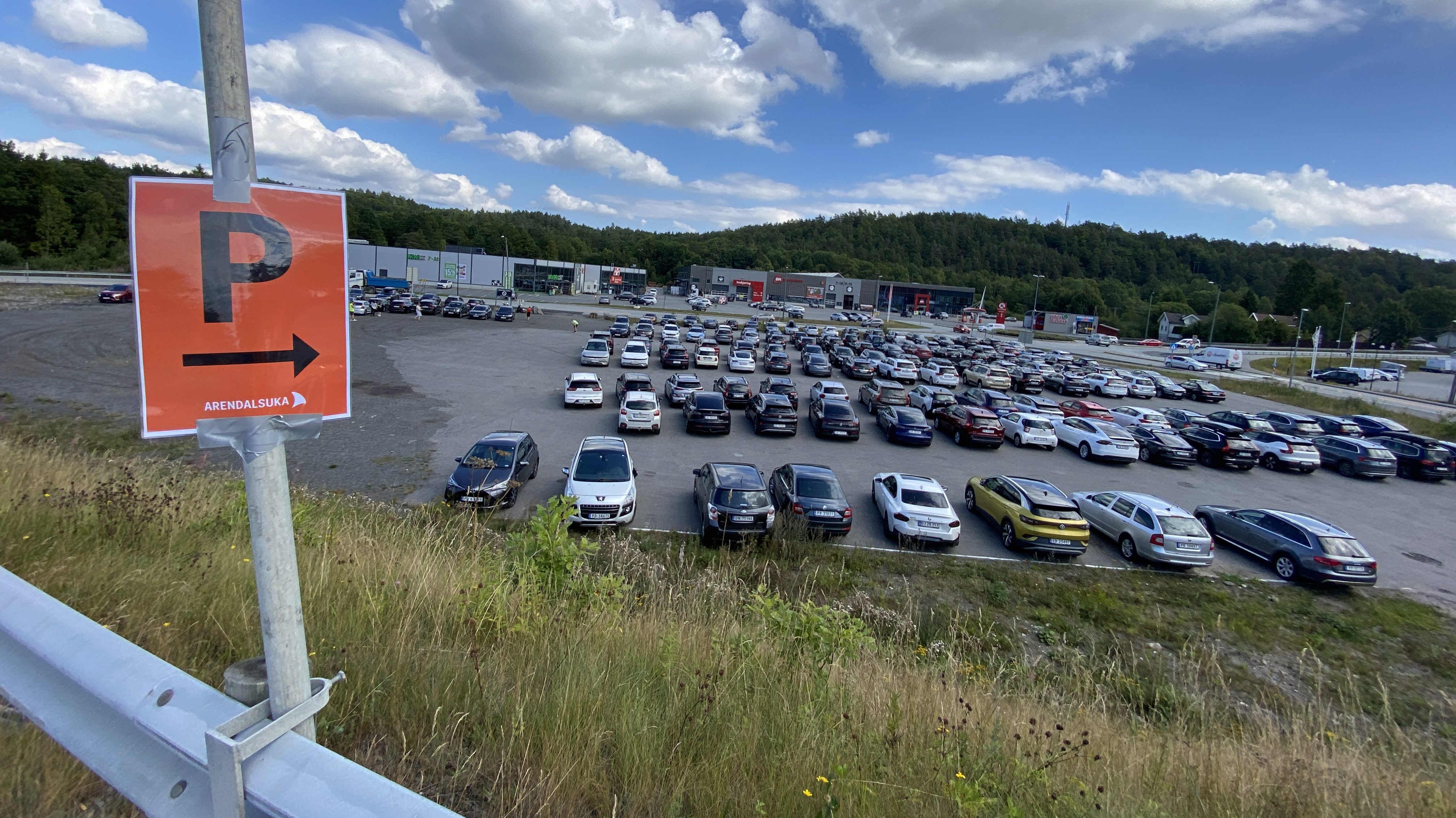 Et vidvinkelbilde av en stor, utendørs parkeringsplass fylt med mange biler under en blå himmel med hvite skyer. I forgrunnen til venstre står et oransje skilt festet til en stolpe med gaffateip. Skiltet viser en stor bokstav "P", en pil som peker til høyre, og teksten "ARENDALSUKA" nederst. Parkeringsplassen ligger i et flatt område med næringsbygg i bakgrunnen, omkranset av skogkledde åser.