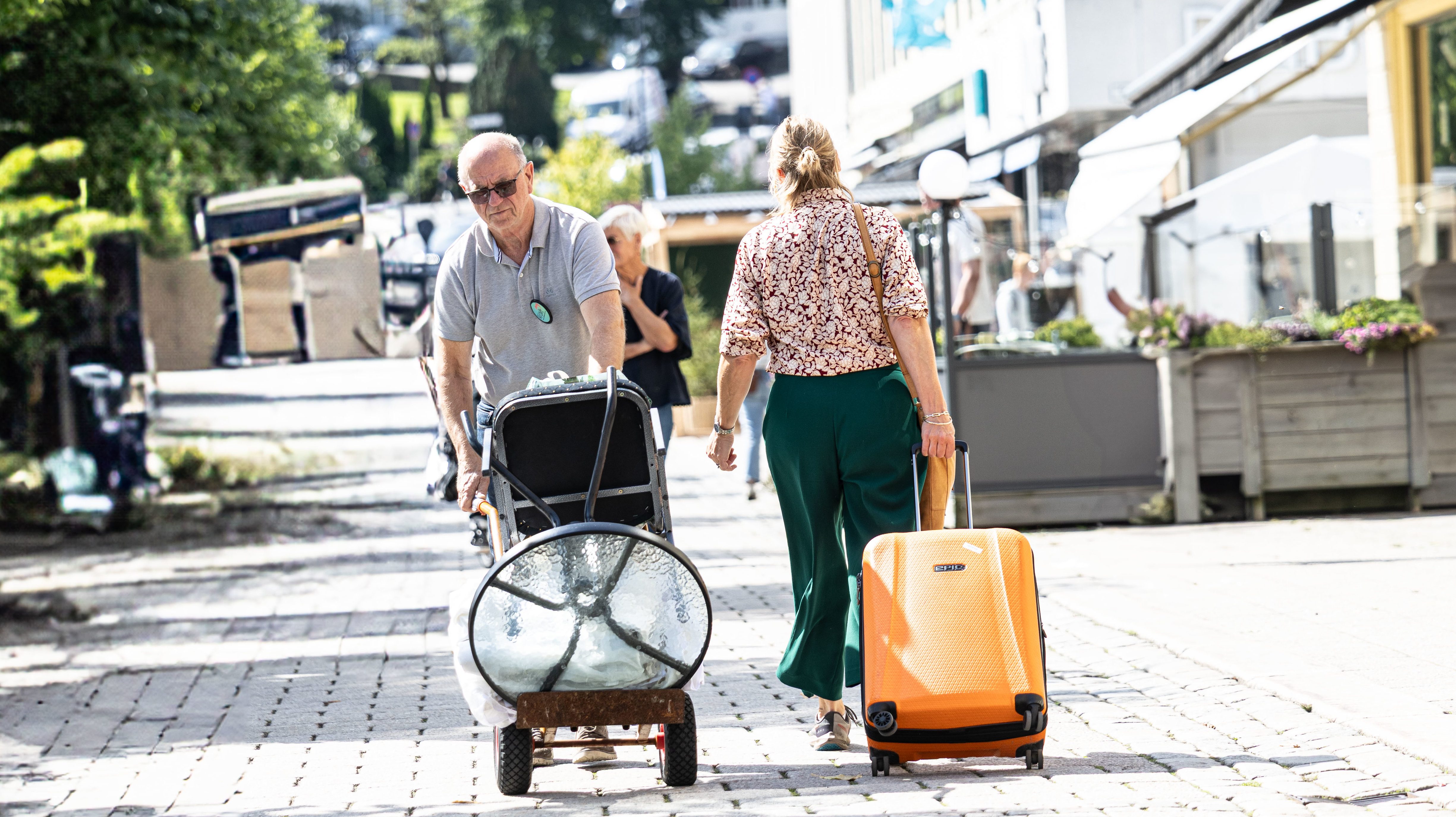 En mann og en kvinne beveger seg bortover en brosteinsgate i dagslys. Mannen til venstre triller en håndtralle lastet med et rundt glassbord og en stol. Kvinnen til høyre går ved siden av ham og drar på en stor, oransje koffert. I bakgrunnen ses hvite bygårder, trær og andre fotgjengere under en lys himmel.