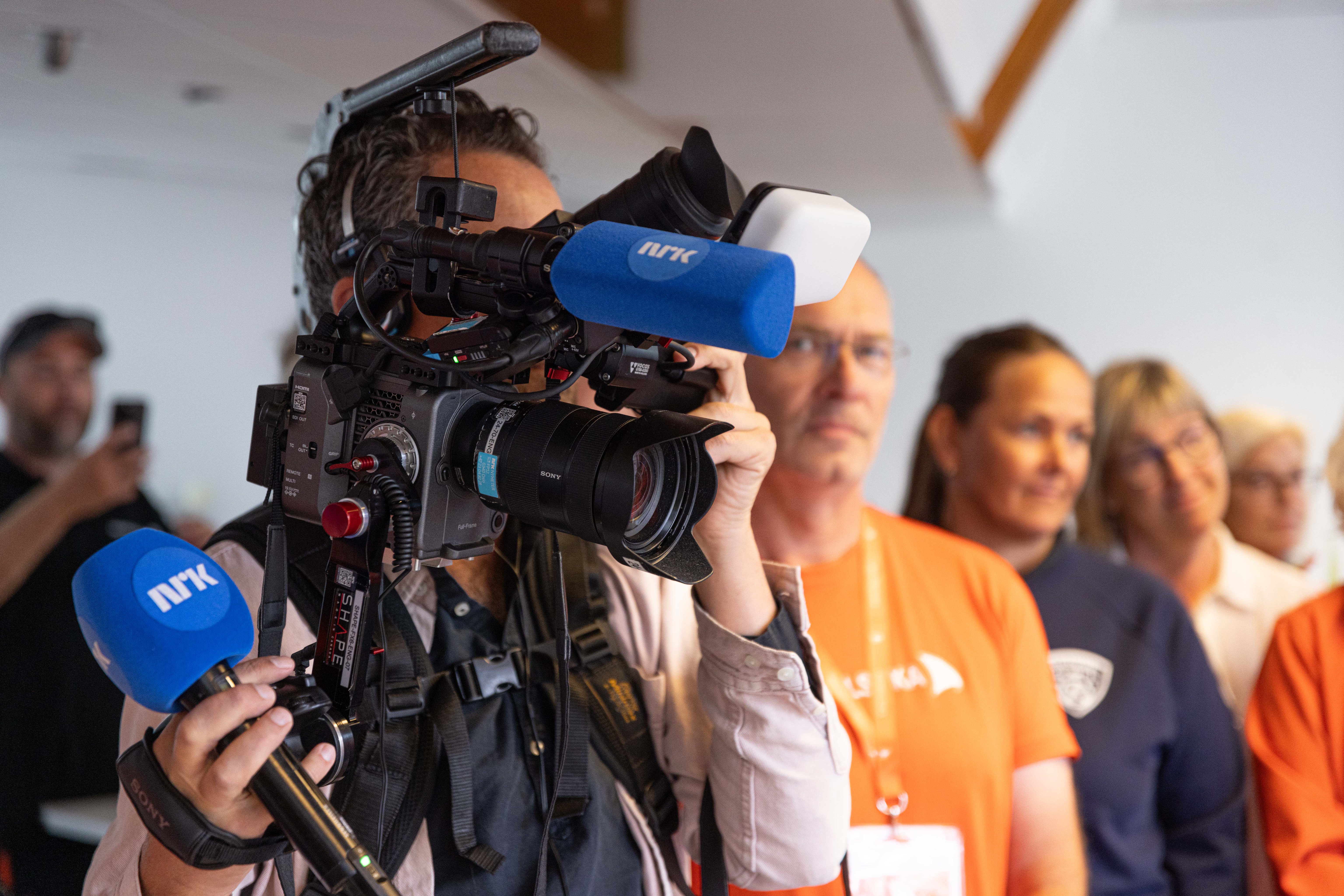 En nærbilde av en fotograf i profil som holder et stort profesjonelt videokamera med NRK-logo på mikrofonene. Fotografen bærer kameraet på skulderen ved hjelp av en bæresele. I bakgrunnen står flere personer på rekke og ser spent på noe utenfor bildet, inkludert en mann i en oransje t-skjorte med "Arendalsuka"-logo. Bakgrunnen er lys og litt ute av fokus, noe som setter alt fokuset på medieutstyret i forgrunnen.