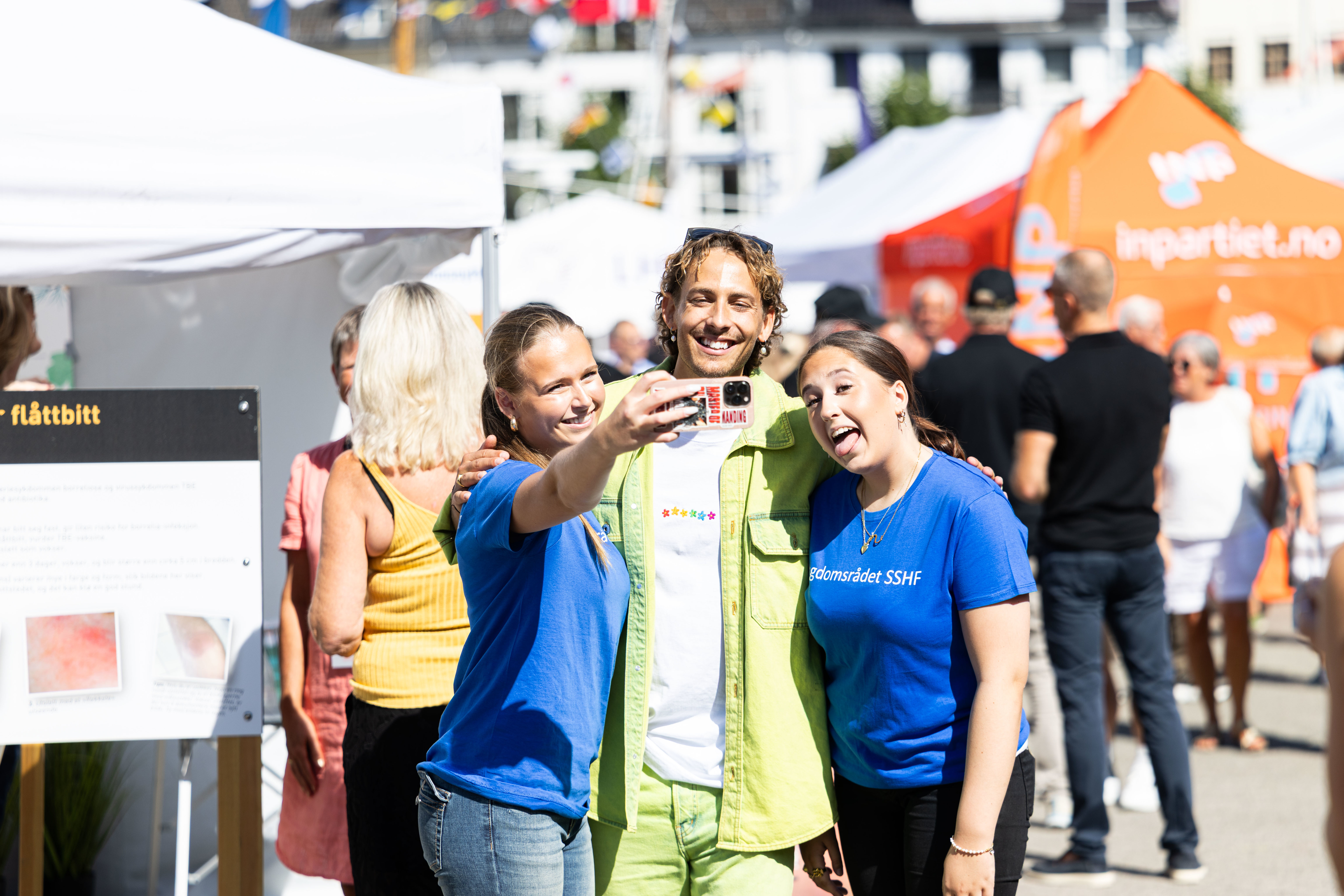 Tre smilende unge mennesker tar en selfie sammen på en solfylt dag under Arendalsuka. En person i limegrønn skjorte står i midten med armene rundt to kvinner i blå t-skjorter merket med "Ungdomsrådet SSHF". I bakgrunnen er det et travelt folkeliv med hvite og oransje partitelt, samt en informasjonsplakat om flåttbitt. Stemningen er munter og uformell midt i arrangementet.