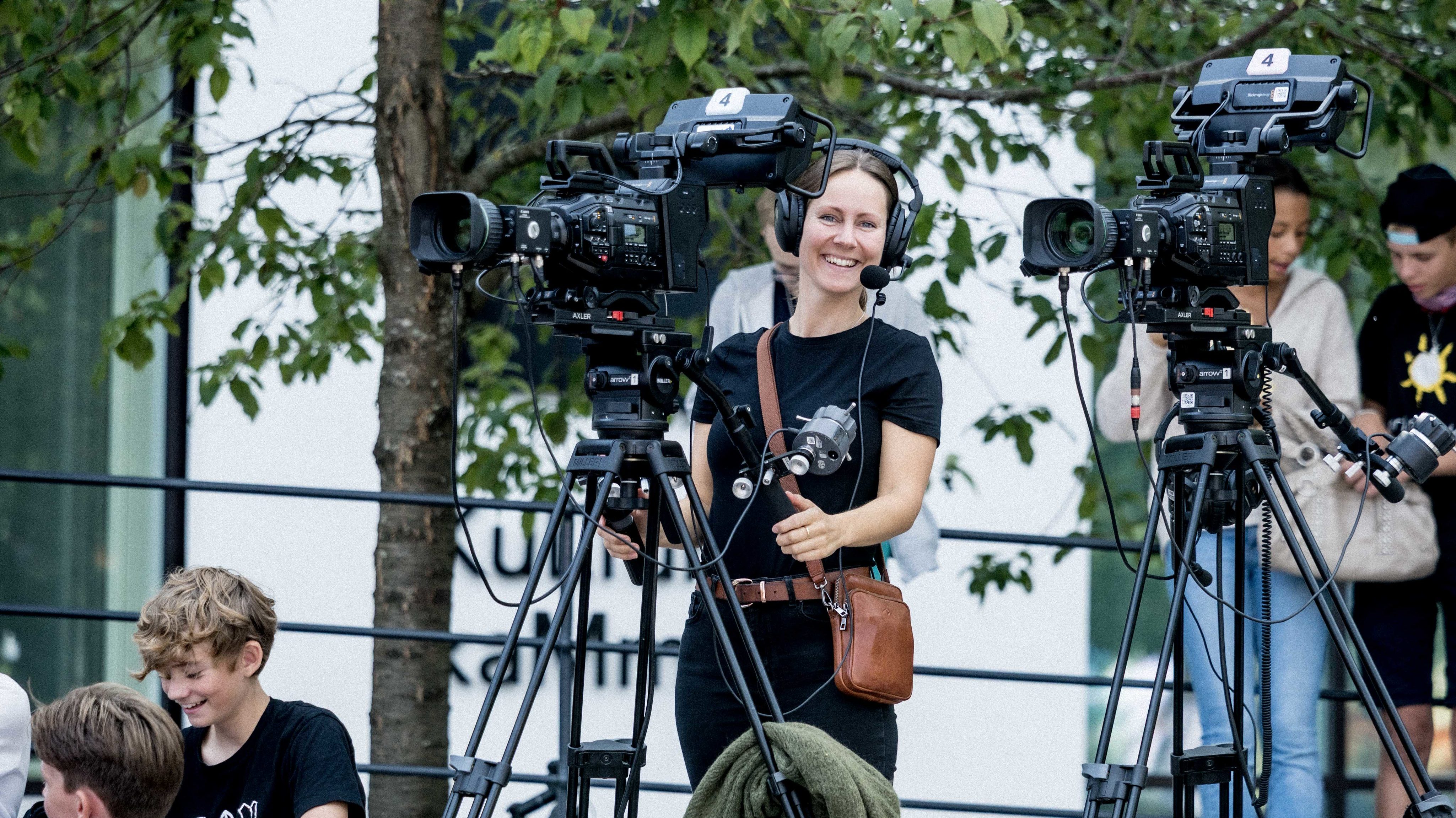 En smilende kvinne med hodetelefoner og mikrofon står bak et profesjonelt filmkamera montert på et stativ utendørs. Hun er kledd i svart og har en brun skulderveske. Ved siden av henne står et tilsvarende kamera, også rettet mot et publikum som sitter på bakken i forgrunnen. I bakgrunnen ses grønne trær og en hvit bygning med store vinduer, noe som indikerer en profesjonell strømming eller TV-produksjon fra et arrangement under Arendalsuka.