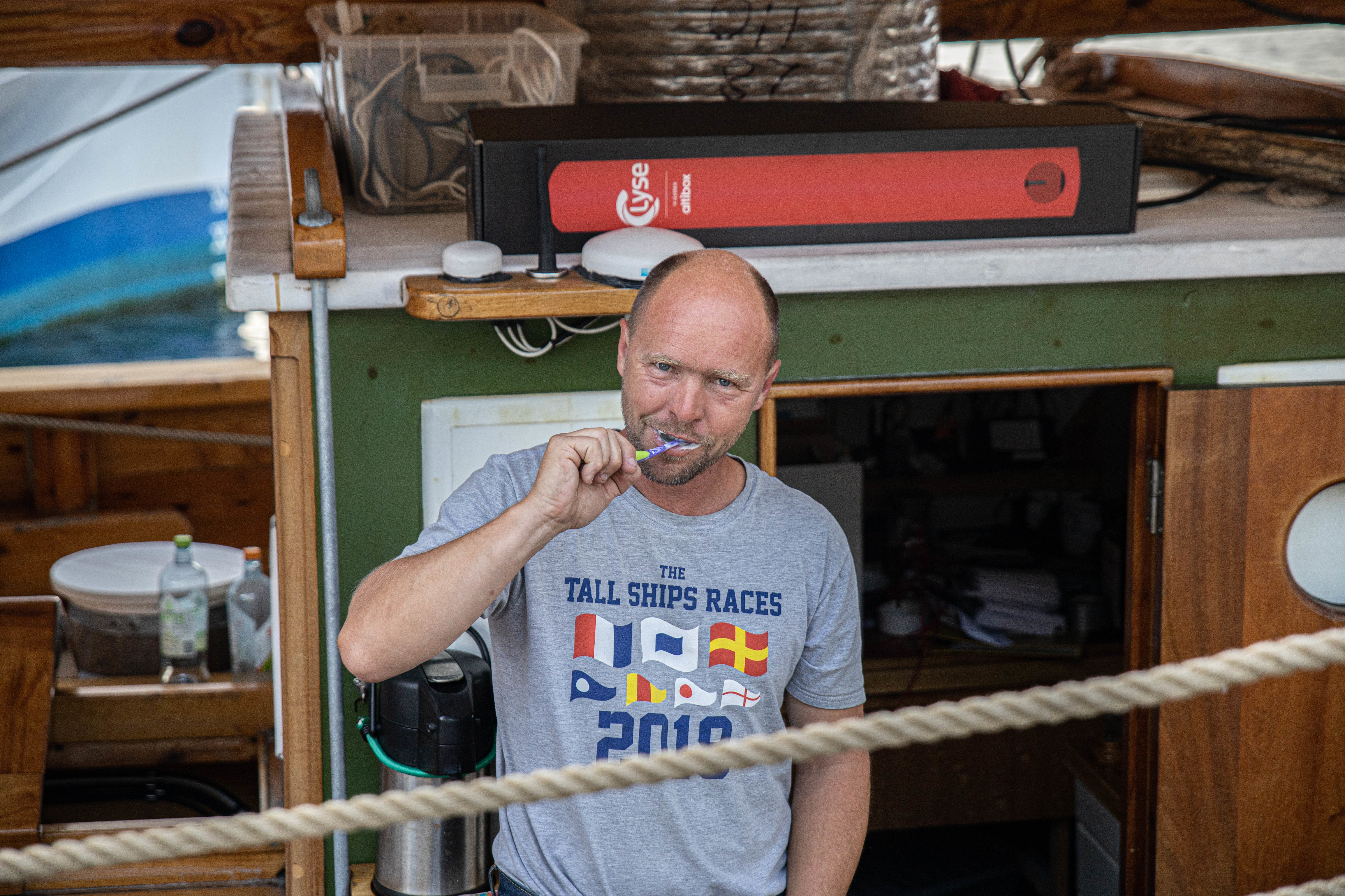En mann står om bord i en trebåt og pusser tennene mens han ser direkte inn i kamera. Han har på seg en grå t-skjorte med teksten "The Tall Ships Races 2019" og illustrasjoner av maritime signalflagg. I bakgrunnen ser man detaljer fra båtens dekk, inkludert en grønnmalt vegg, en rød boks merket med "Lyse", og diverse utstyr. Et tykt tau løper diagonalt i forgrunnen av bildet.