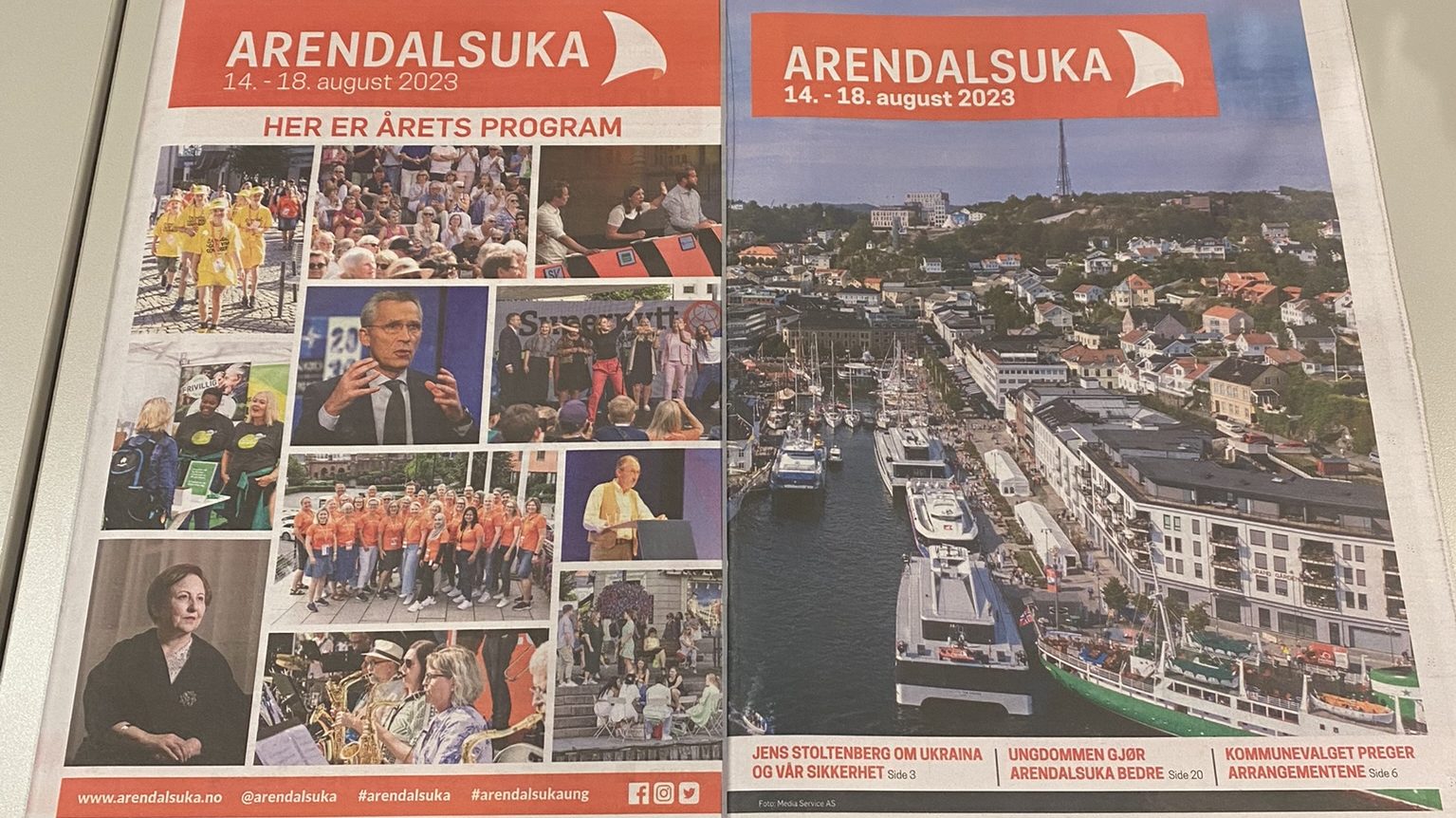 Et trykt programhefte for Arendalsuka 2023 ligger oppslått på et grått skrivebord ved siden av en PC-skjerm. Venstresiden av heftet viser en kollasj med små bilder av talere, publikum, frivillige og konserter under overskriften "Her er årets program". Høyresiden viser et stort oversiktsbilde av Pollen i Arendal fylt med båter og folk, med overskriften "Arendalsuka 14.–18. august 2023" på en oransje bakgrunn. Nederst på siden er det henvisninger til artikler om blant annet Jens Stoltenberg og ungdommens rolle.