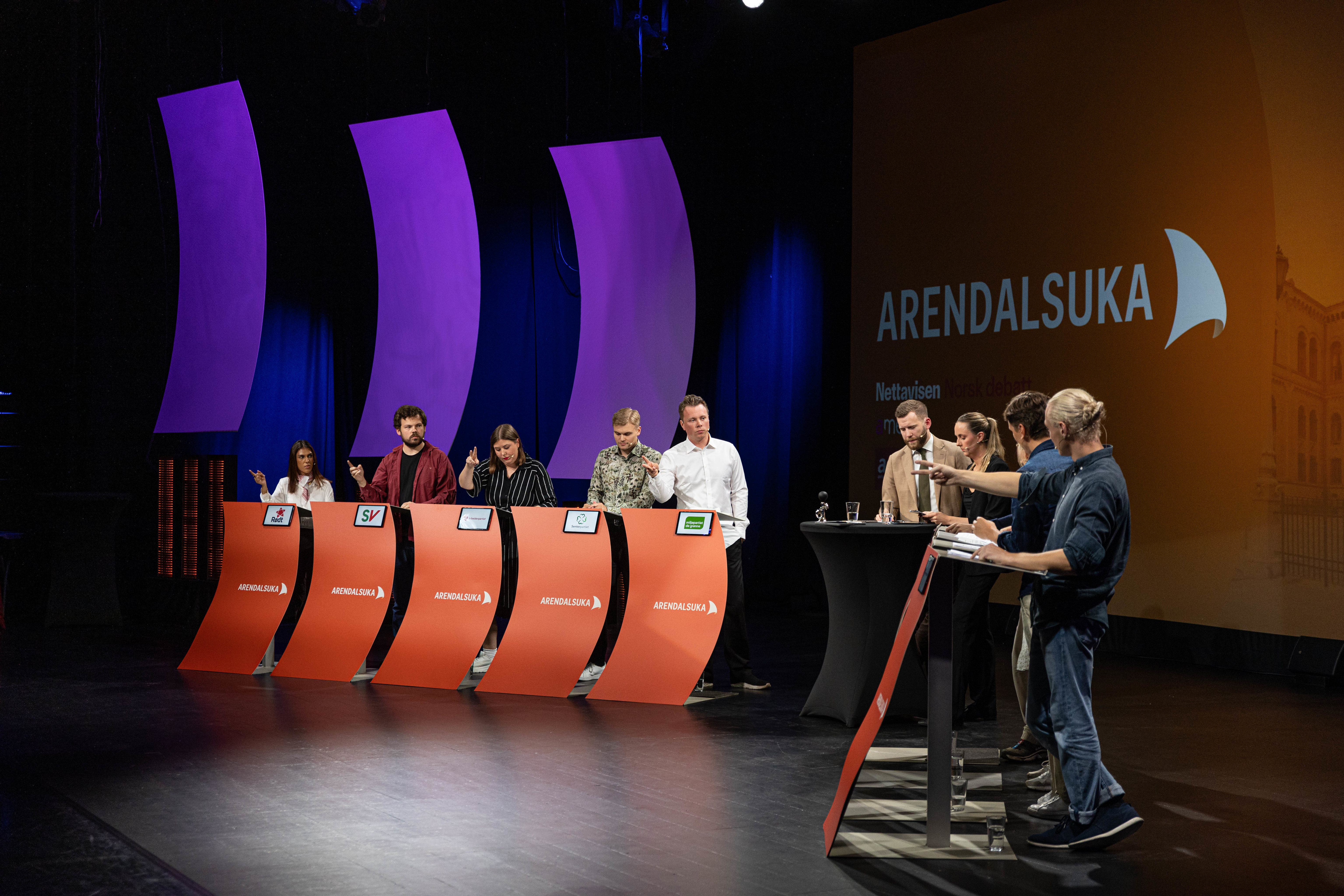 En politisk debatt på en mørk scene under Arendalsuka. Fem debattanter står bak hver sin buede, oransje talerstol med Arendalsuka-logoen. Talerstolene er merket med logoene til Rødt, SV, Arbeiderpartiet, Senterpartiet og Miljøpartiet De Grønne. Til høyre står to moderatorer ved et rundt bord, og teknisk personell er synlig i forkant. Bak debattantene er det tre store, lilla seil-lignende dekorasjoner, og en stor skjerm til høyre viser teksten "ARENDALSUKA" over et bilde av en historisk bygning.
