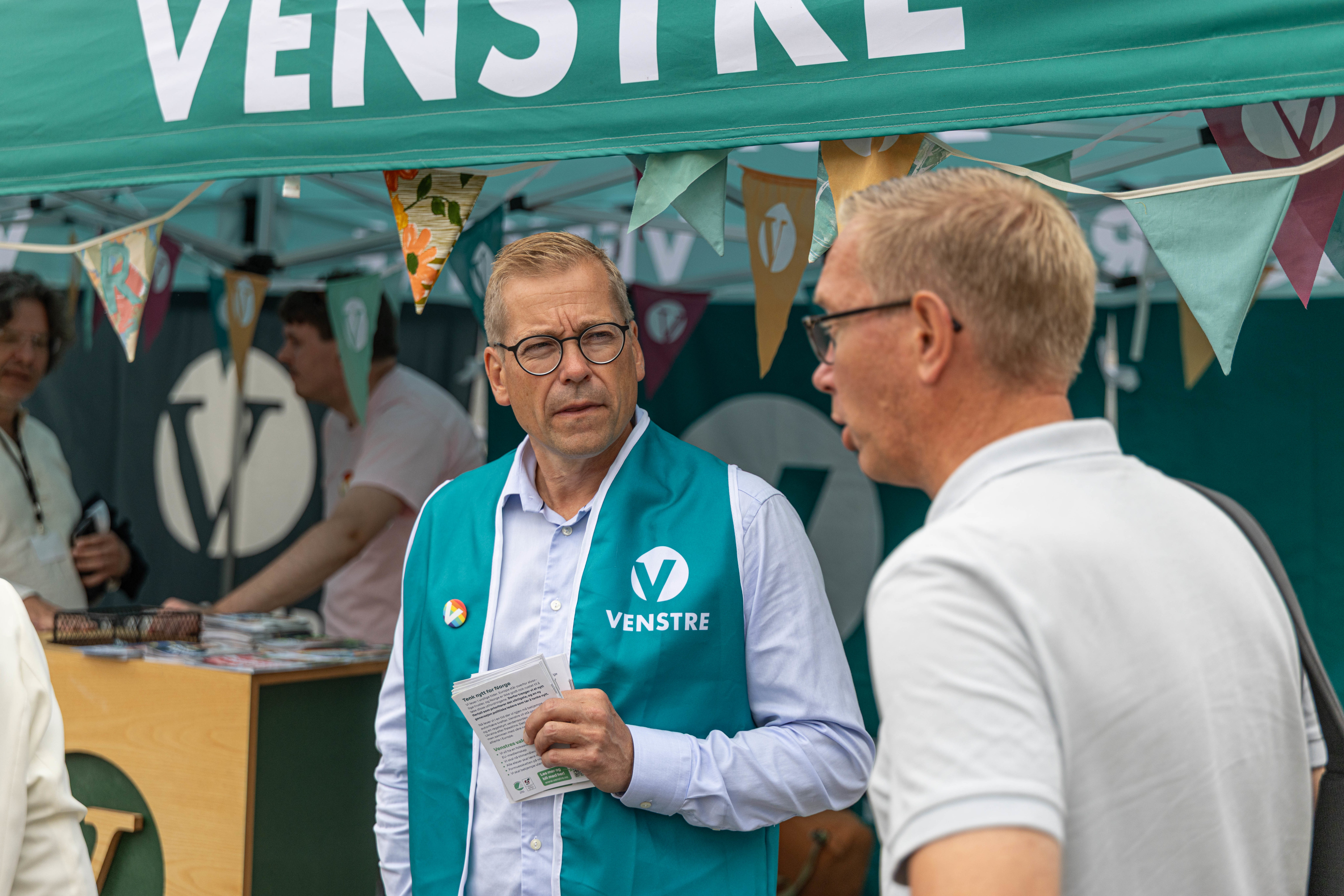 To menn står i profil og snakker sammen under en grønn teltduk merket med "VENSTRE" i store, hvite bokstaver. Mannen til venstre har på seg en grønn Venstre-vest over en lyseblå skjorte, bærer briller og holder en bunke brosjyrer. Teltet er pyntet med små vimpelflagg i ulike farger og mønstre. I bakgrunnen kan man se en annen person og en disk med informasjonsmateriell, noe som skaper en typisk stands-atmosfære under Arendalsuka.