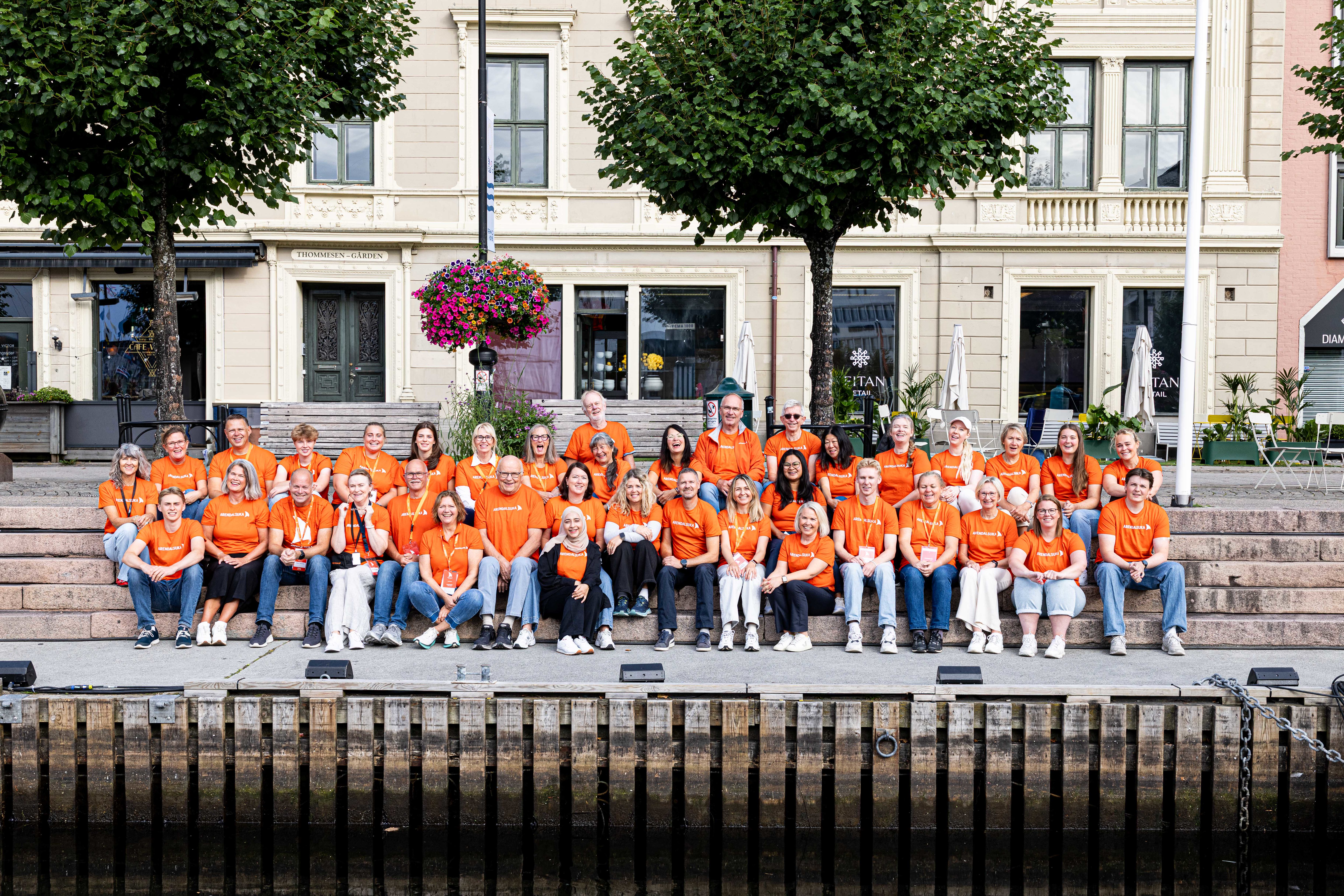 Et stort gruppebilde av vertskapet for Arendalsuka, bestående av cirka 40 personer i alle aldre som sitter og står i flere rekker på en steintrapp ved bryggekanten i Pollen. Alle er kledd i like oransje t-skjorter med Arendalsuka-logoen. Bak dem ses historiske bygårder, trær og blomsterkasser under en klar himmel. I forgrunnen speiler bryggekanten seg i det stille vannet.