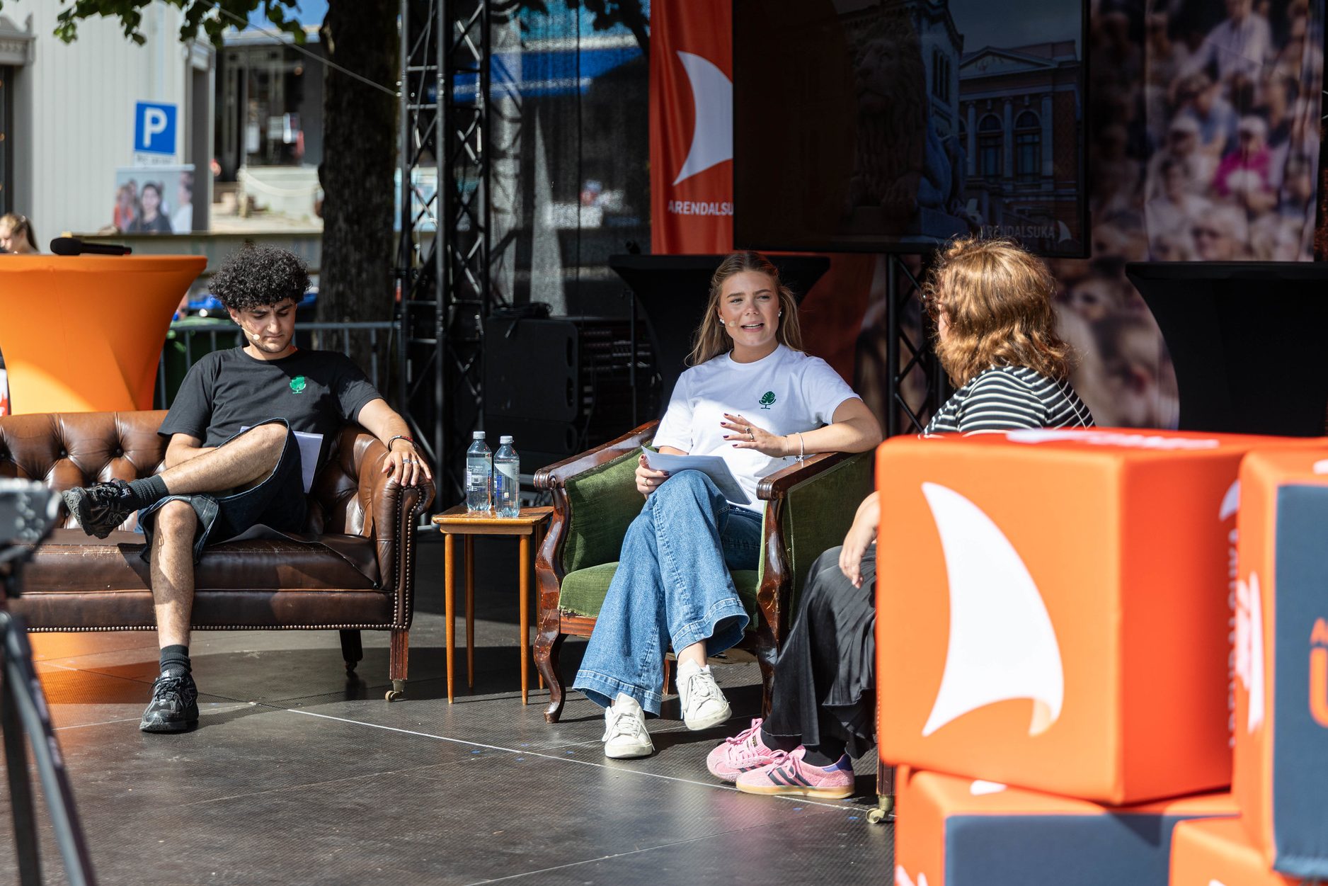 Tre personer sitter i en samtale i en sofa og lenestoler på en utendørsscene. I forgrunnen står flere oransje sittekuber merket med logoen til Arendalsuka. En TV-skjerm og grønne trær er synlig i bakgrunnen under arrangementet.
