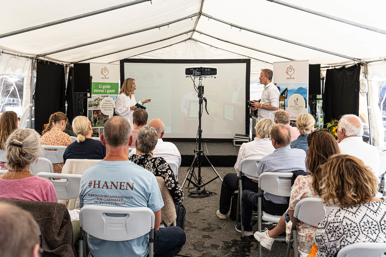 To personer presenterer foran et lerret i et fullsatt arrangementstelt under Arendalsuka. Publikum sitter på hvite klappstoler og følger med på scenen.