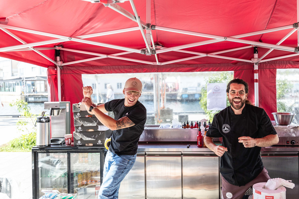To smilende ansatte i en matbod under Arendalsuka. De står bak en rustfri ståldisk under et rødt telttak, med Pollens havnebasseng synlig i bakgrunnen.