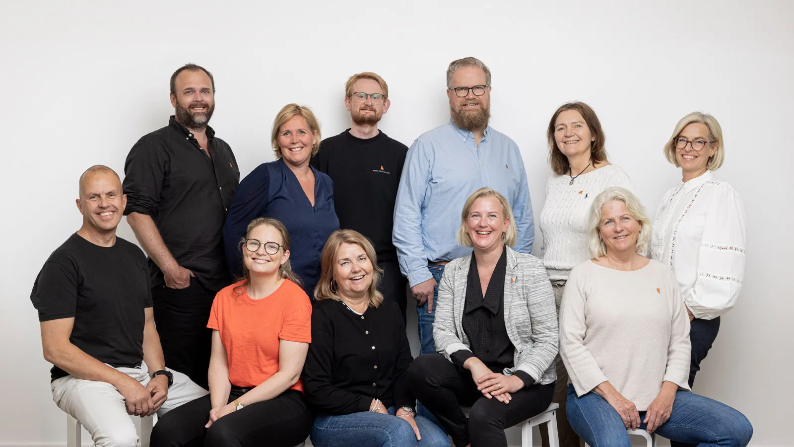 Et gruppebilde av Arendalsukas prosjektgruppe - elleve smilende personer foran en hvit vegg. De er kledd i hverdagslige klær, og flere har på seg små oransje pins med Arendalsuka-logoen.