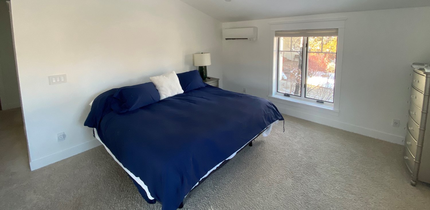 A bright, minimalist bedroom with a large bed dressed in blue bedding, a window letting in natural light, and a wall-mounted mini-split unit above the window