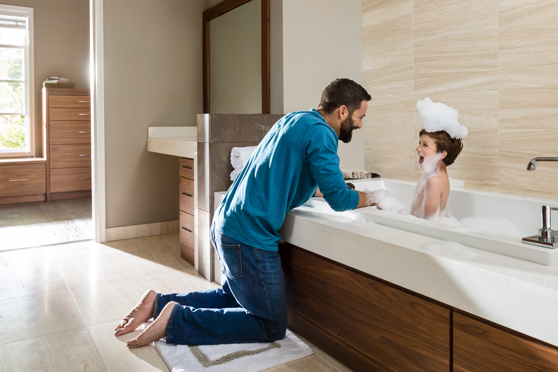A parent kneeling beside a bathtub, smiling and playing with a laughing child covered in bubbles during bath time in a bright, modern bathroom
