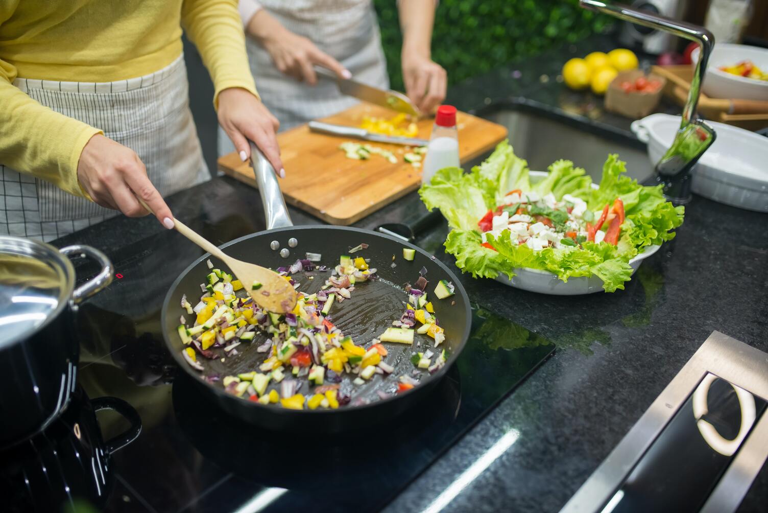Two people preparing vegetables in a kitchen, with one sautéing chopped vegetables in a pan on an induction stovetop and another chopping ingredients nearby