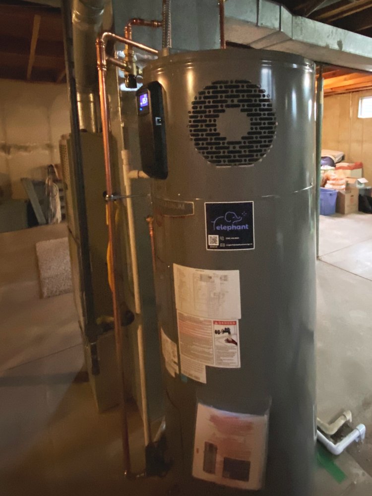 “A heat pump water heater installed in a basement, with copper piping, ventilation components, and an Elephant Energy sticker visible on the unit