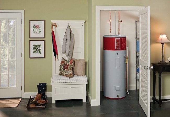A heat pump water heater installed in a small utility closet off a hallway, with copper piping visible and nearby household storage and decor