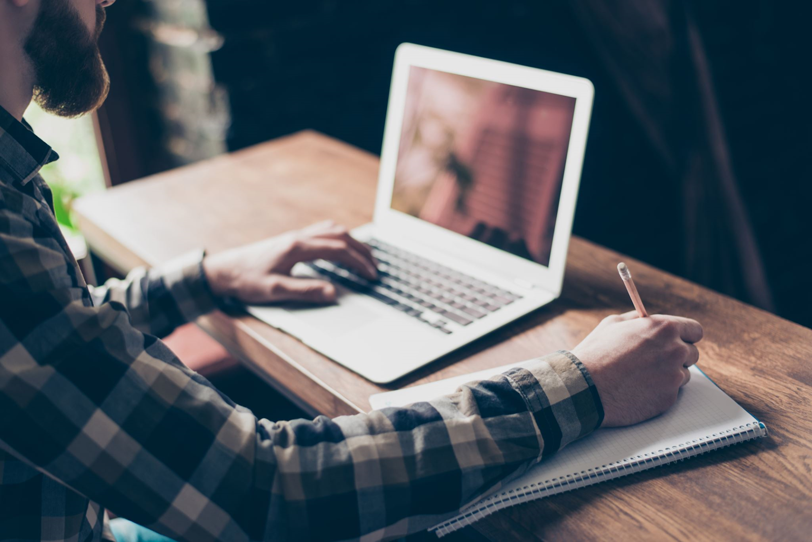 A person sitting at a wooden desk, writing a 360 plan in a notebook while working on a laptop in a softly lit indoor setting