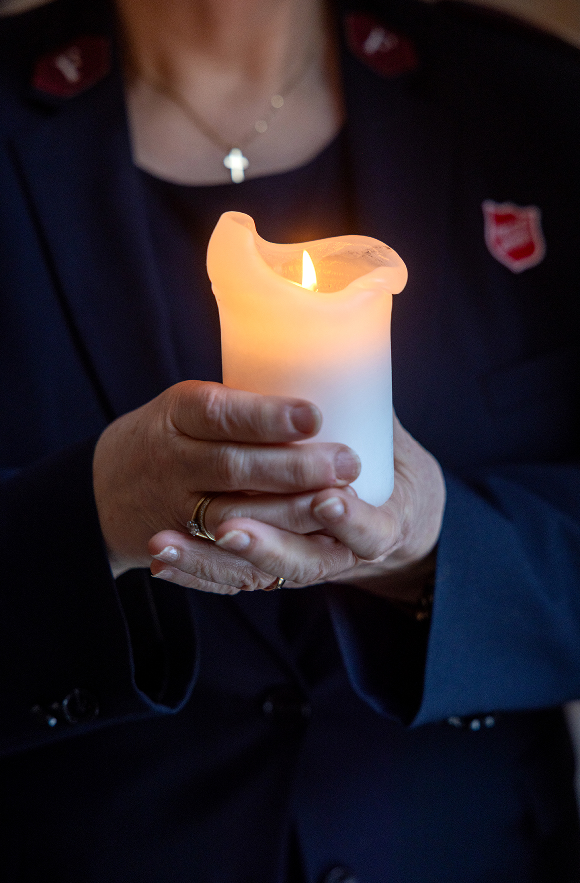 Foto: Nærbilde av to hender som holder et kubbelys. Personen synes ikke, men man kan se en mørk jakke med Frelsesarme-skjold uskarpt bak lyset.
