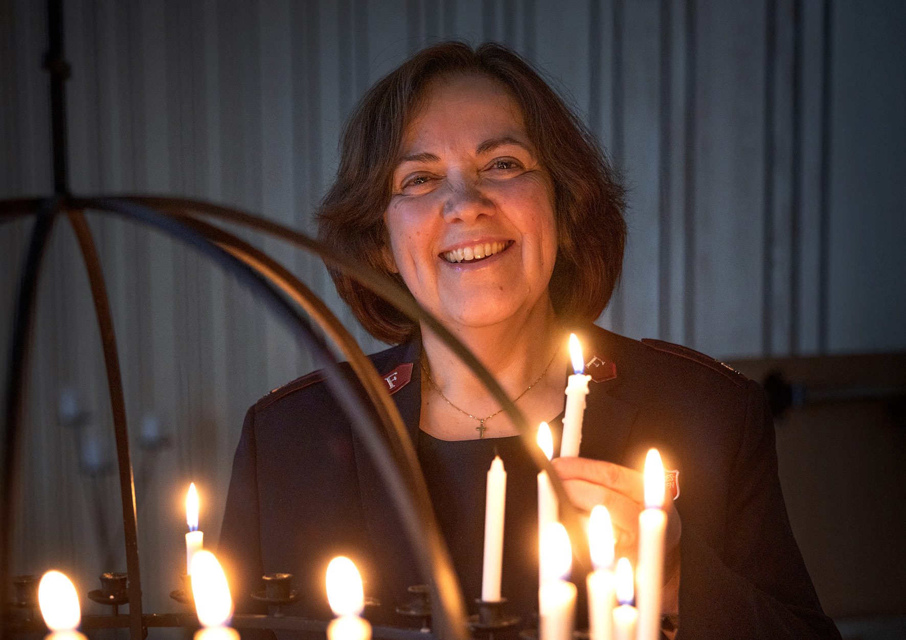Foto: En kvinne med halvlangt, brunt hår står og smiler mot kamera. Bare ansikt og overkropp er med på bildet. Hun holder et tent stearinlys i hånden, og foran henne vises deler av en lysglobe med tente lys.