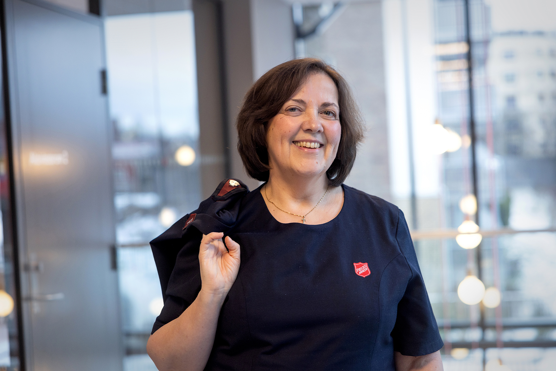 Foto: Bilde i halvfigur av en kvinne med mørkt halvlangt hår som smiler mot kamera. Hun holder en jakke over den ene skulderen. I bakgrunnen sees en åpen dør og store vinduer, som er uskarpe på bildet.