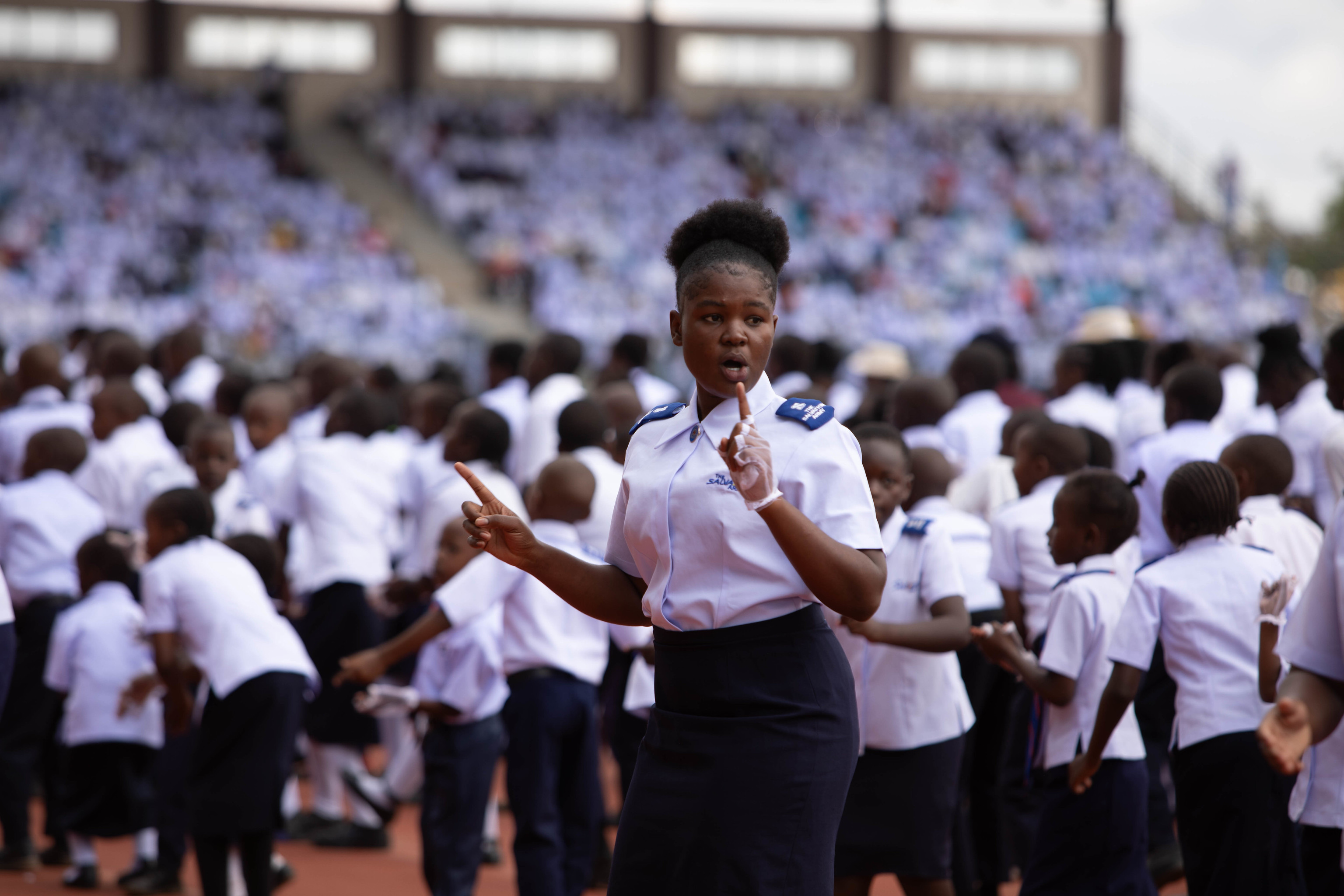 4 bilder: 1. Generalen i hvit uniform står på talerstolen på et utendørs stadion. 2. Ung afrikansk kvinnelig soldat danser foran mange andre unge soldater. 3. Hornorkester i røde jakker marsjer inn på stadion, Kenyas og Frelsesarmeens flagg bæres foran. Leder og nestleder med koner, alle i hvite uniformer marsjerere inn på stadion