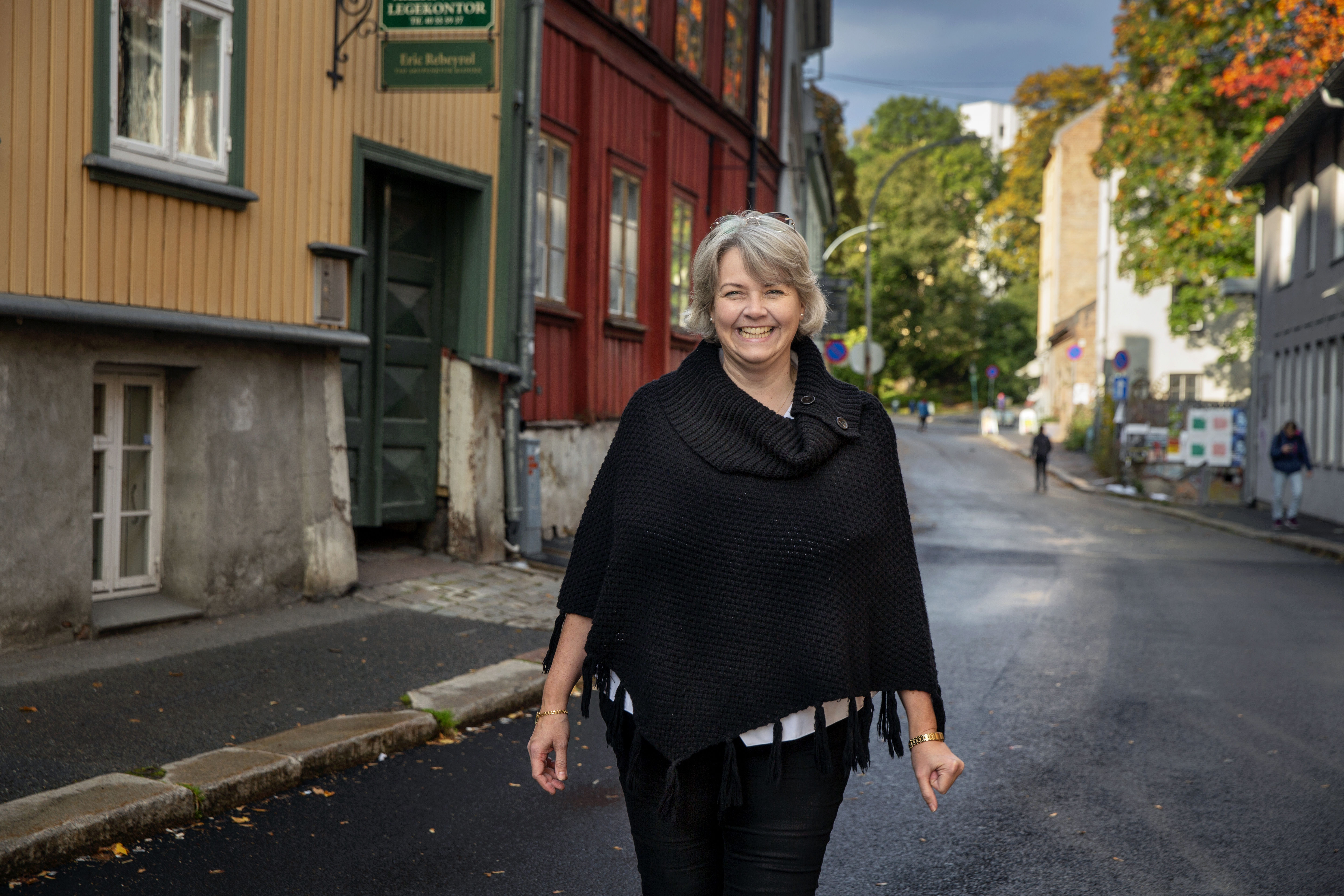 En smilende dame går mot kamera. i Bakgrunnen ser vi en Oslogate med trehus i friske farger.