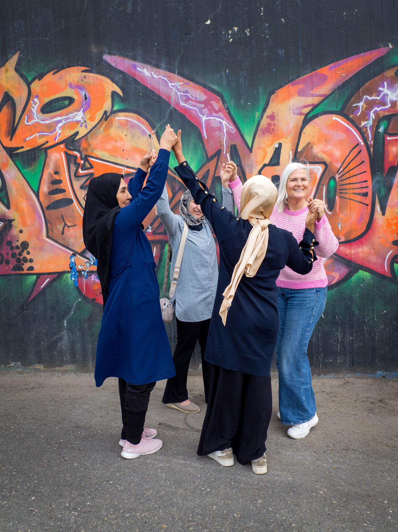 Foto: En norsk dame med hvitt hår og rosa og hvit-stripete genser holder hender og går i ring med tre damer med hijab.