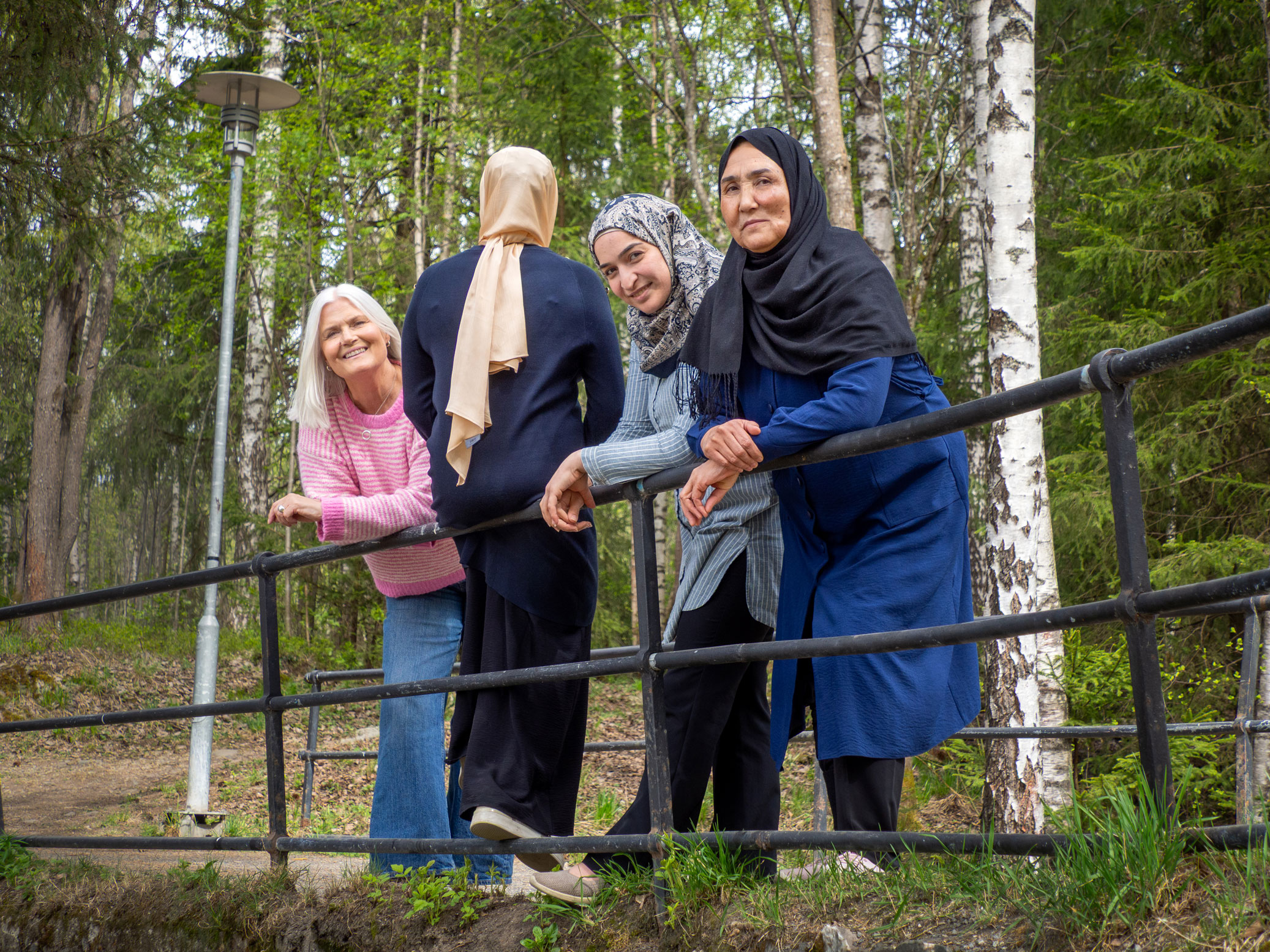 Foto: En norsk kvinne med grått hår og rosa og hvit-stripete ullgenser lener seg ut over et rekkverk i smijern ute i naturen. Hun står sammen med tre kvinner med hijab, der den ene ikke viser sitt ansikt.