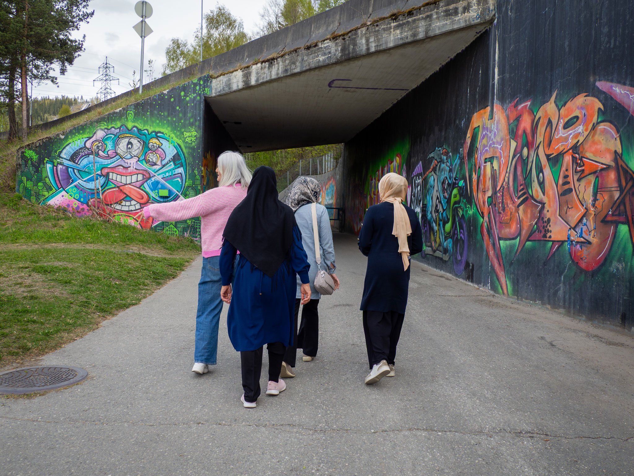 Foto: Fire kvinner går på tur innover mot en undergang med taggin på begge sider av betongen. Kvinnen med hvitt hår i rosa og hvit-stripene ullgenser peker bort på gresset. De andre tre har hijab.