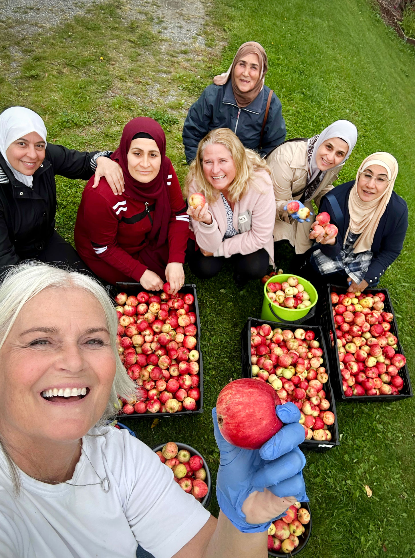 Foto: En smilende kvinne med grått hår holder opp en rødt eple. På selfien hun tar er det seks andre kvinner i bakgrunnen, fem med hijab, som sitter foran flere kasser med epler.