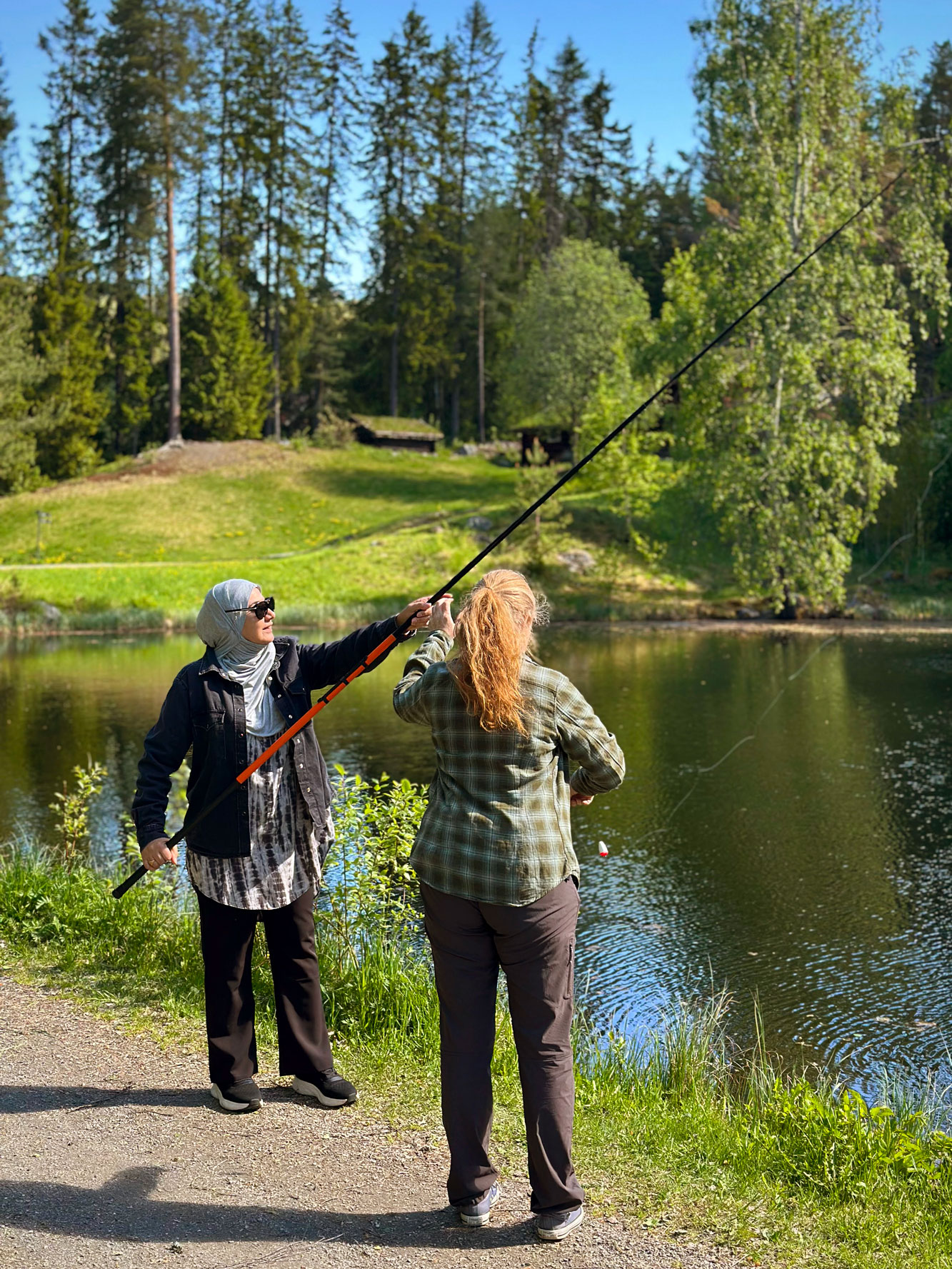 Foto: Kvinne med rødt, langt hår hjelper en kvinne med hijab. Hun holder opp en fiskestang og de står foran et vann.