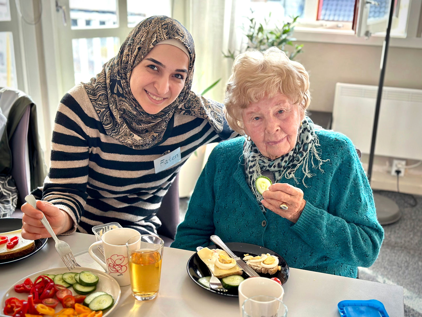 Foto: Yngre dame med hijab sitter sammen med en norsk dame på over 80 år. Hun spiser en agurkskive.